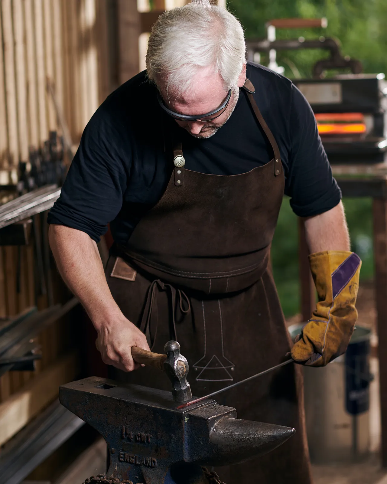 A blacksmith in a brown apron is hammering the red-hot end of an iron rod into a sharp point on an anvil.