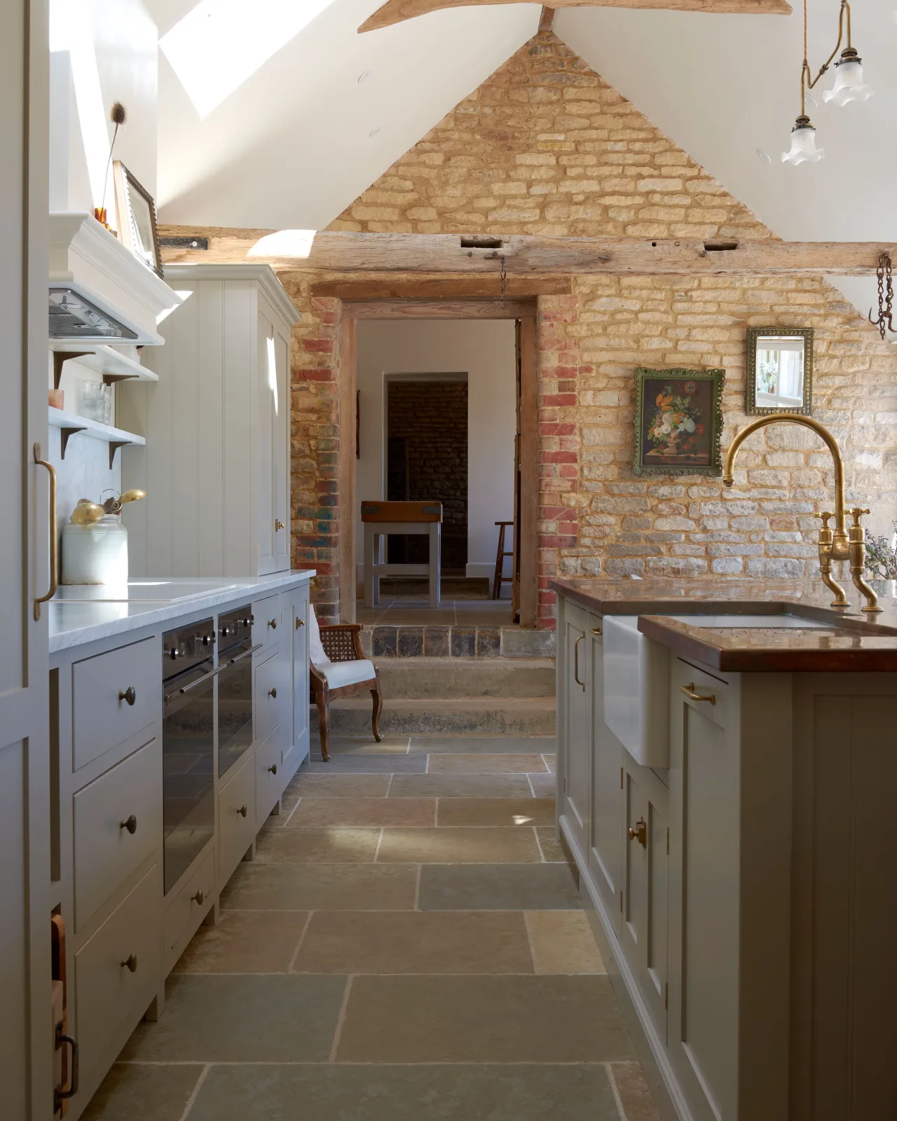 A view looking between a run of Mushroom Shaker kitchen cabinets and a copper-topped Shaker island, towards the original stone walls of a barn conversion kitchen, with exposed wooden beams, glints of Aged Brass and Jaipur Brushed Limestone flooring.