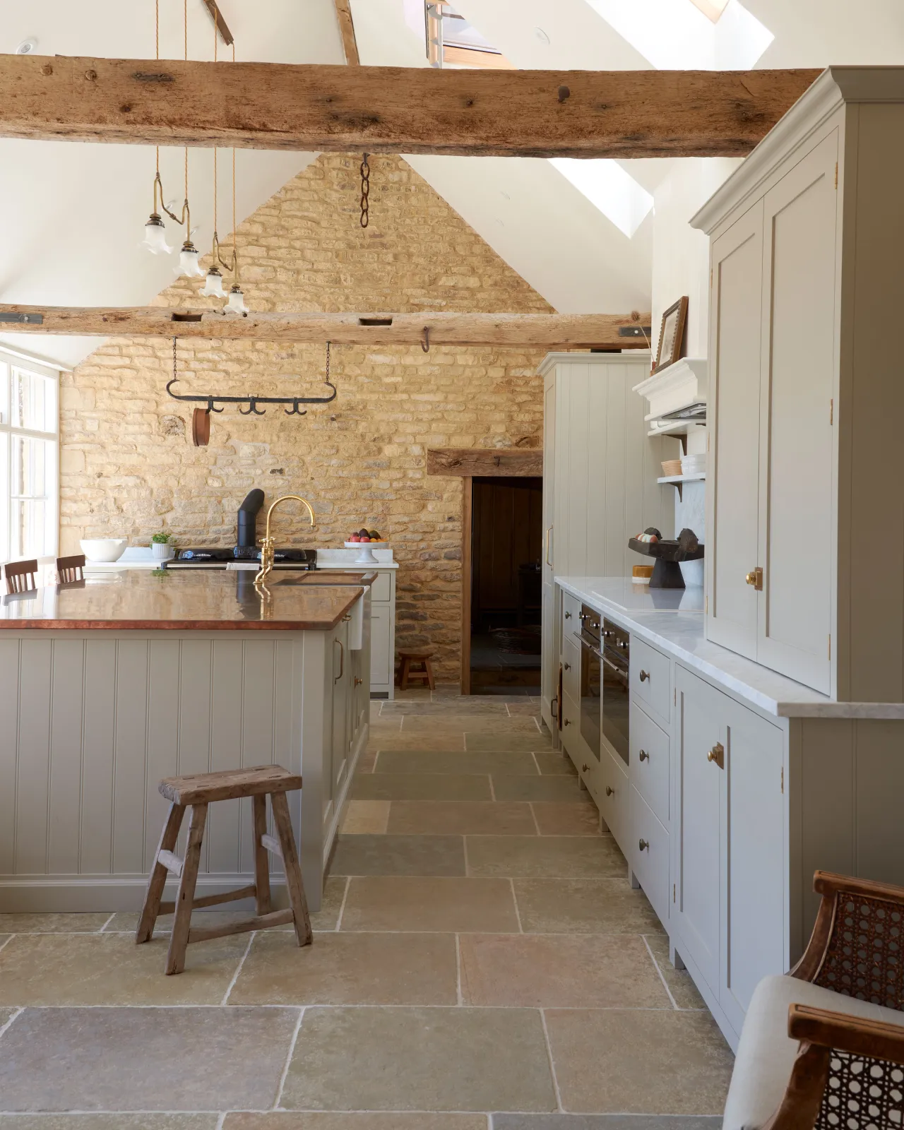 A view looking between a copper-topped Shaker island and a run of Mushroom Shaker kitchen cabinets, towards the original stone walls of a barn conversion kitchen, with exposed wooden beams, glints of Aged Brass and Jaipur Brushed Limestone flooring.