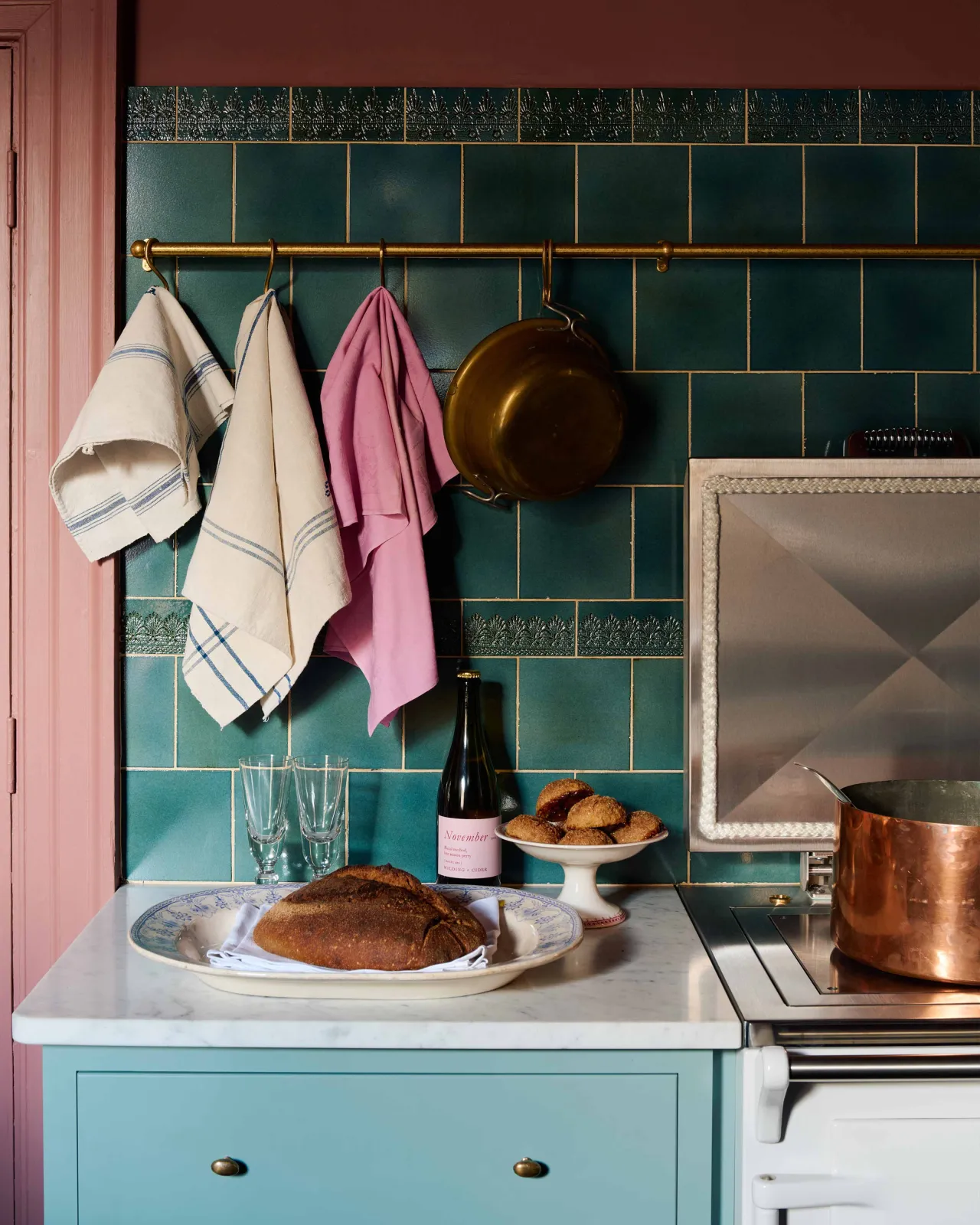 A display of our glossy ‘Lace Market Tiles’ in a rich vintage teal colour, featuring a mix of square tiles in a brickbond pattern and bands of metro tiles with our ‘Flemish’ and ‘Flanders’ pressed lace patterns. The colour of the tiles is complimented by the wine-coloured wall and the ‘Aged Brass Hanging Rail’ which features an assortment of vintage tea towels. A bottle of wine, freshly baked bread, and pastries are in the foreground on the Carrara marble worktop.