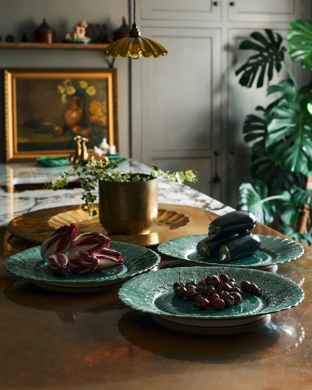 Three large handmade bohemian-style green ceramic platters with an assortement of aubergines, red chicory and cherries in an eclectic kitchen with an aged copper worktop, grey painted walls, antique paintings and a large monstera plant.