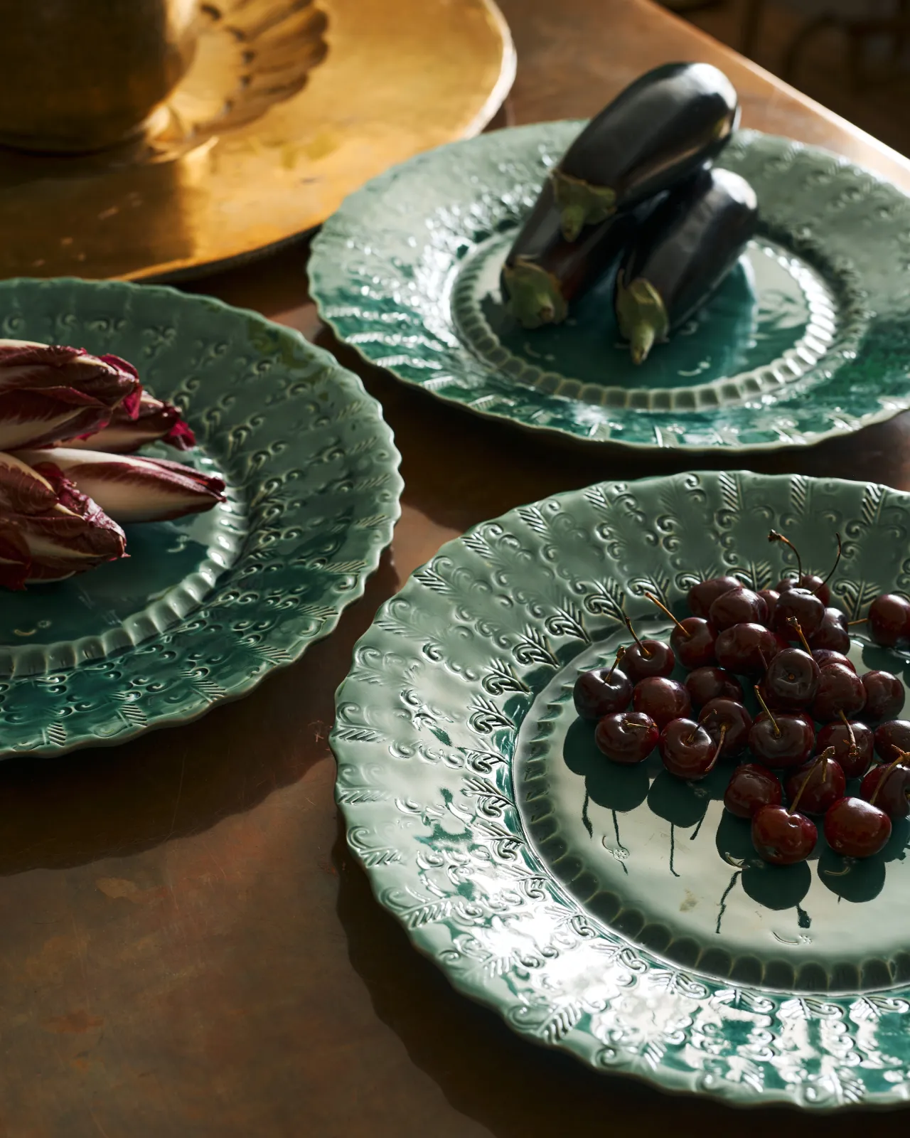 A close-up view of three large handmade bohemian-style green ceramic platters with scalloped rims and delicate impressions and surface patterns and an assortement of aubergines, red chicory and cherries.