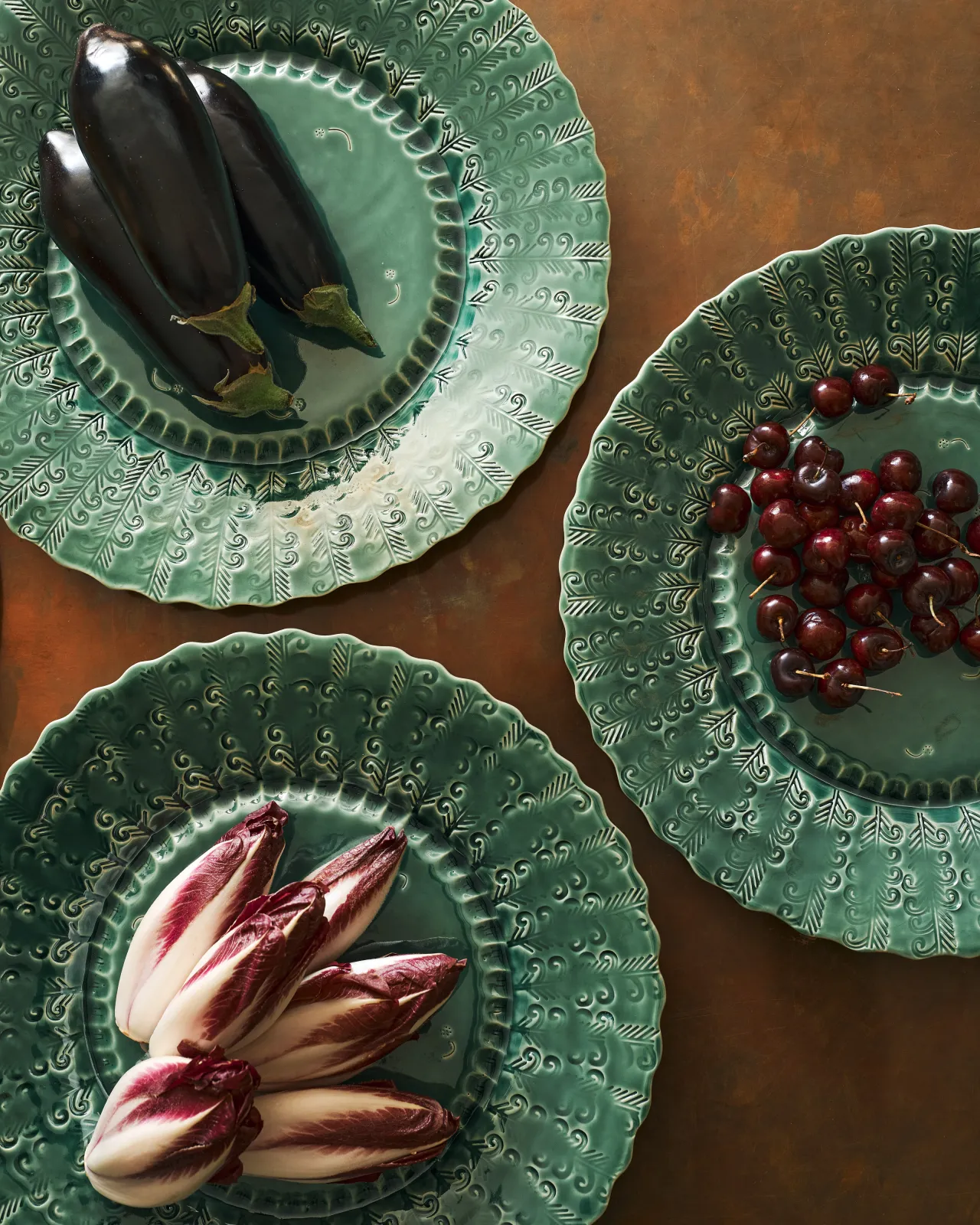 An overhead view of three large handmade bohemian-style green ceramic platters with scalloped rims and delicate impressions and surface patterns and an assortement of aubergines, red chicory and cherries on an aged copper worktop.