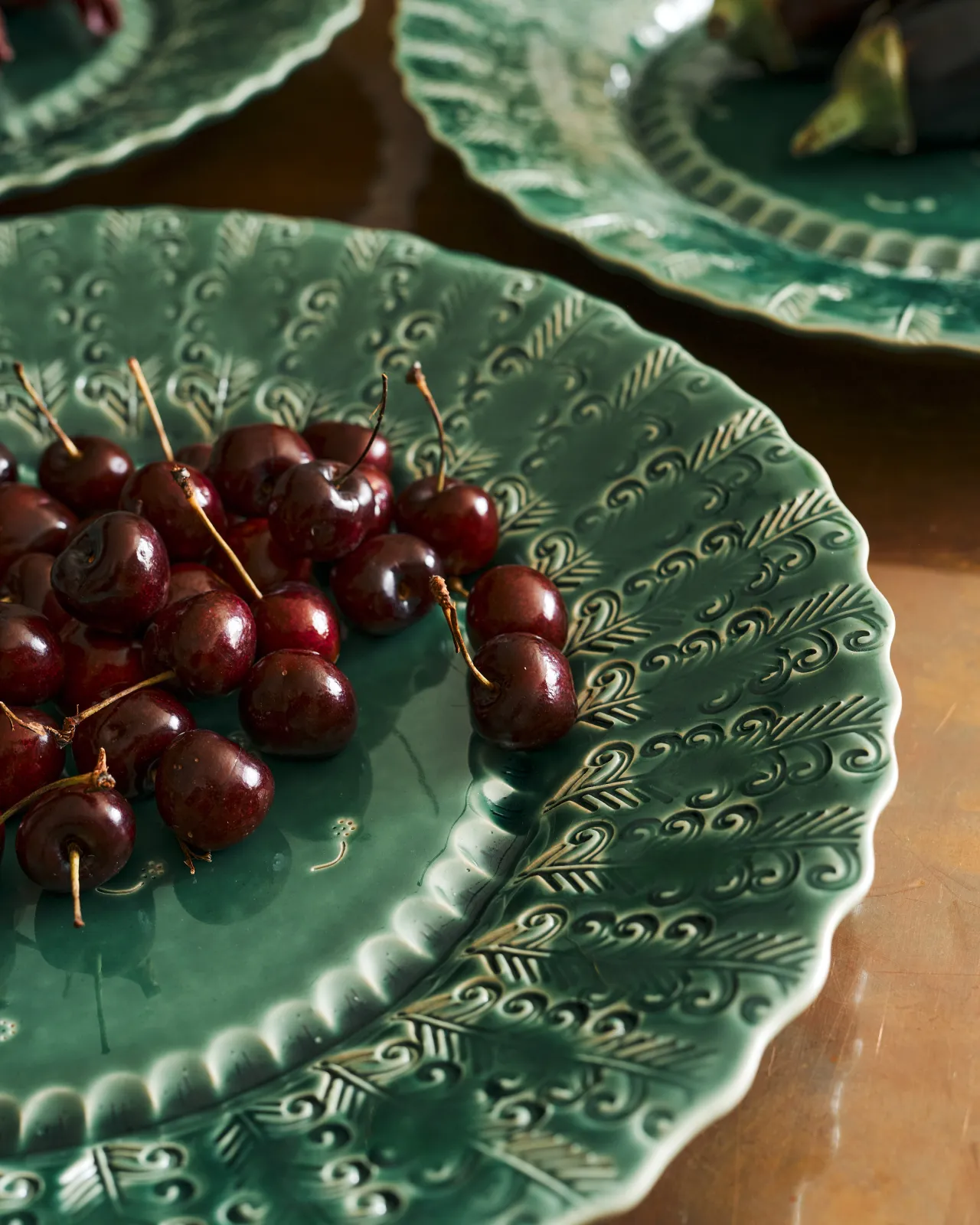 A close-up view of a bunch of cherries on a large handmade bohemian-style green ceramic platter with a scalloped rim and delicate impressions and surface patterns.