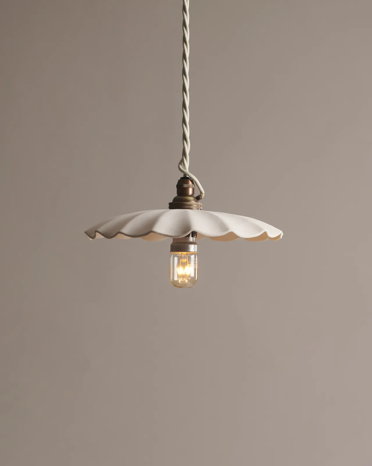 A large wide pleat porcelain pendant light with an unglazed finish, an aged brass B22 lampholder and a white twisted fabric flex cord.