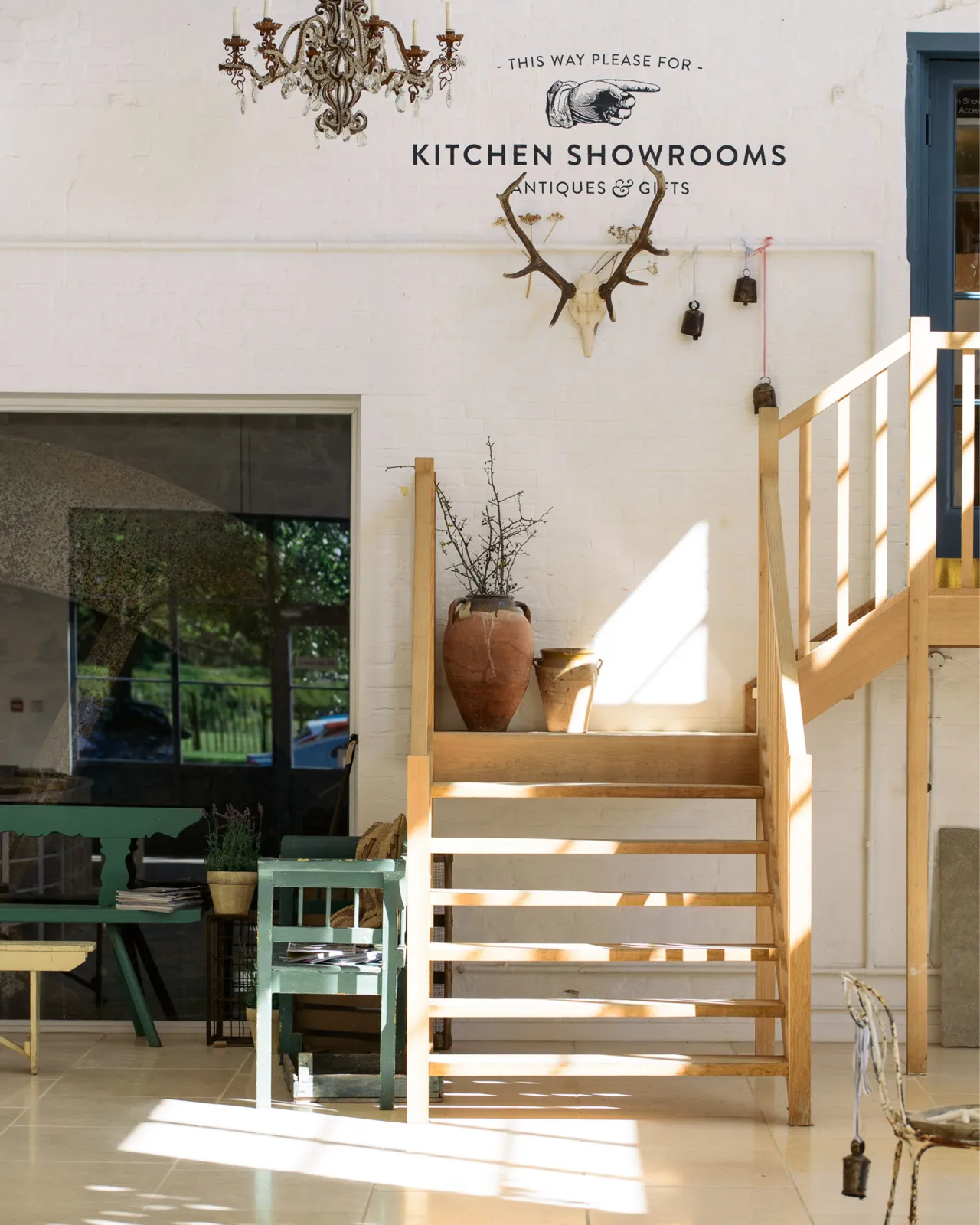 A straight-on shot looking towards the back of deVOL's flooring showroom. Light pours in through the huge glazed windows, casting pools of light and shadows on the wooden staircase and Light Jerusalem Limestone flooring.