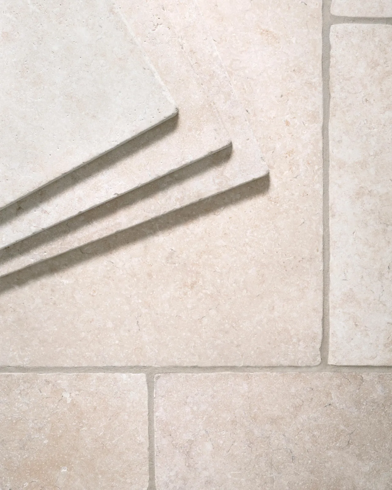A close-up overhead view of Light Jerusalem Limestone flooring, showing the mottled cream colour and smooth texture. Three loose tiles are stacked in the top left corner.