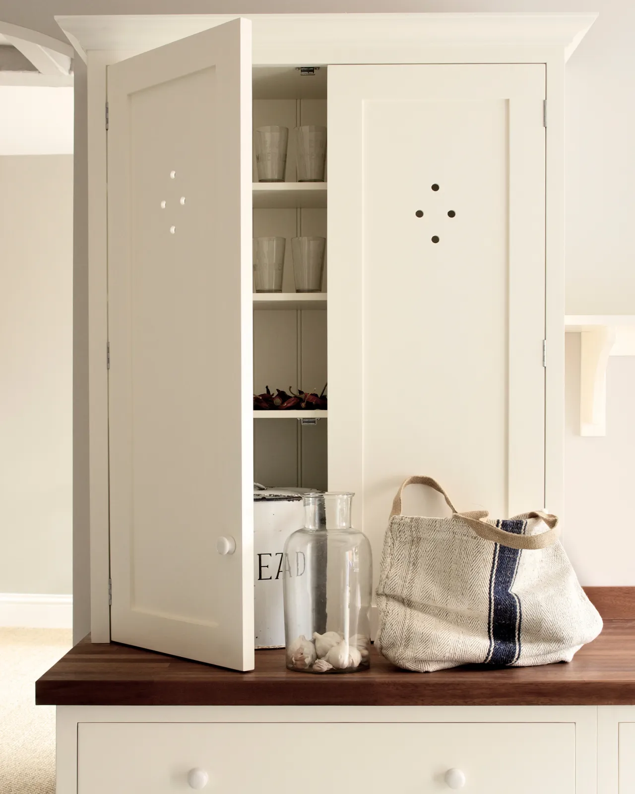 A straight-on view of a simple cream coloured Shaker-style countertop cupboard on a rich hardwood worktop. The door is slightly ajar, revealing a collection of clear glasses inside.