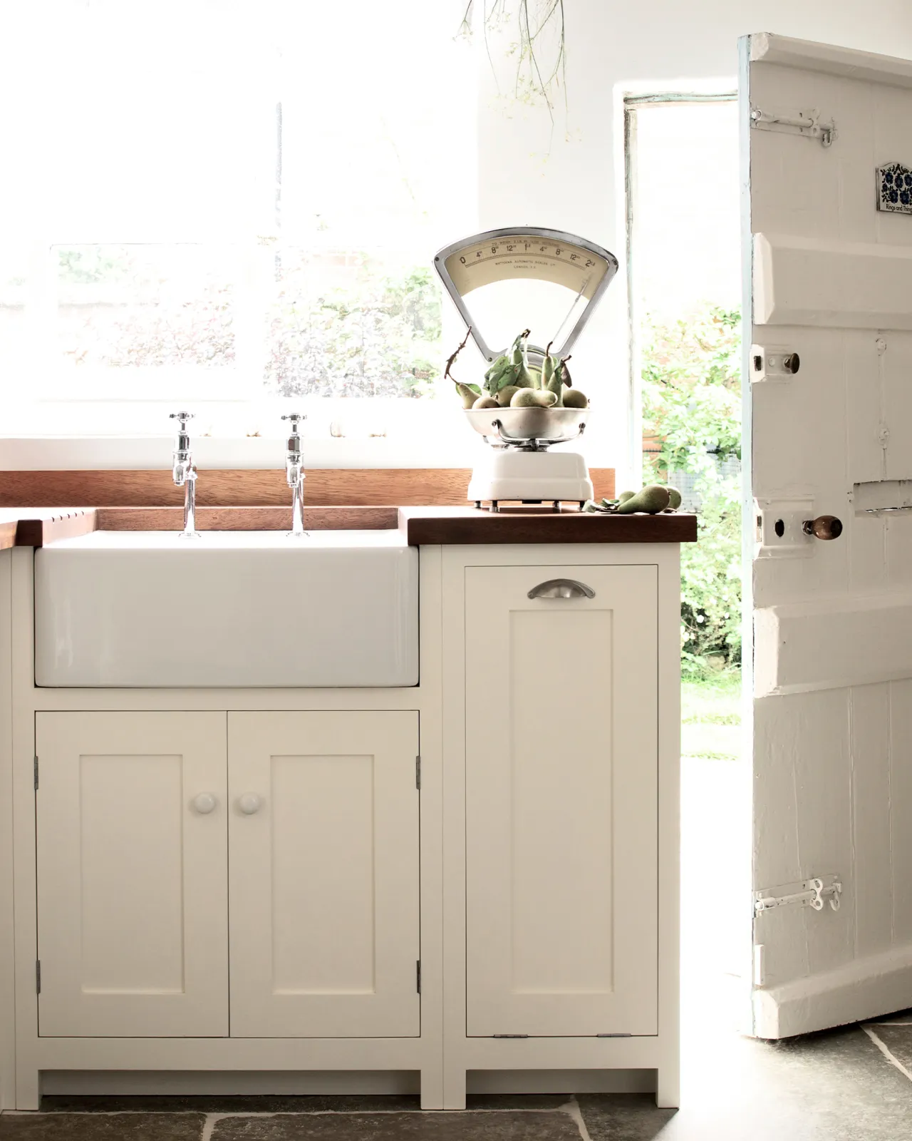 A simple cottage kitchen with cream coloure Shaker-style kitchen cupboards, a rich hardwood worktop, a small Belfast sink and a vintage set of weighing scales on the worktop.