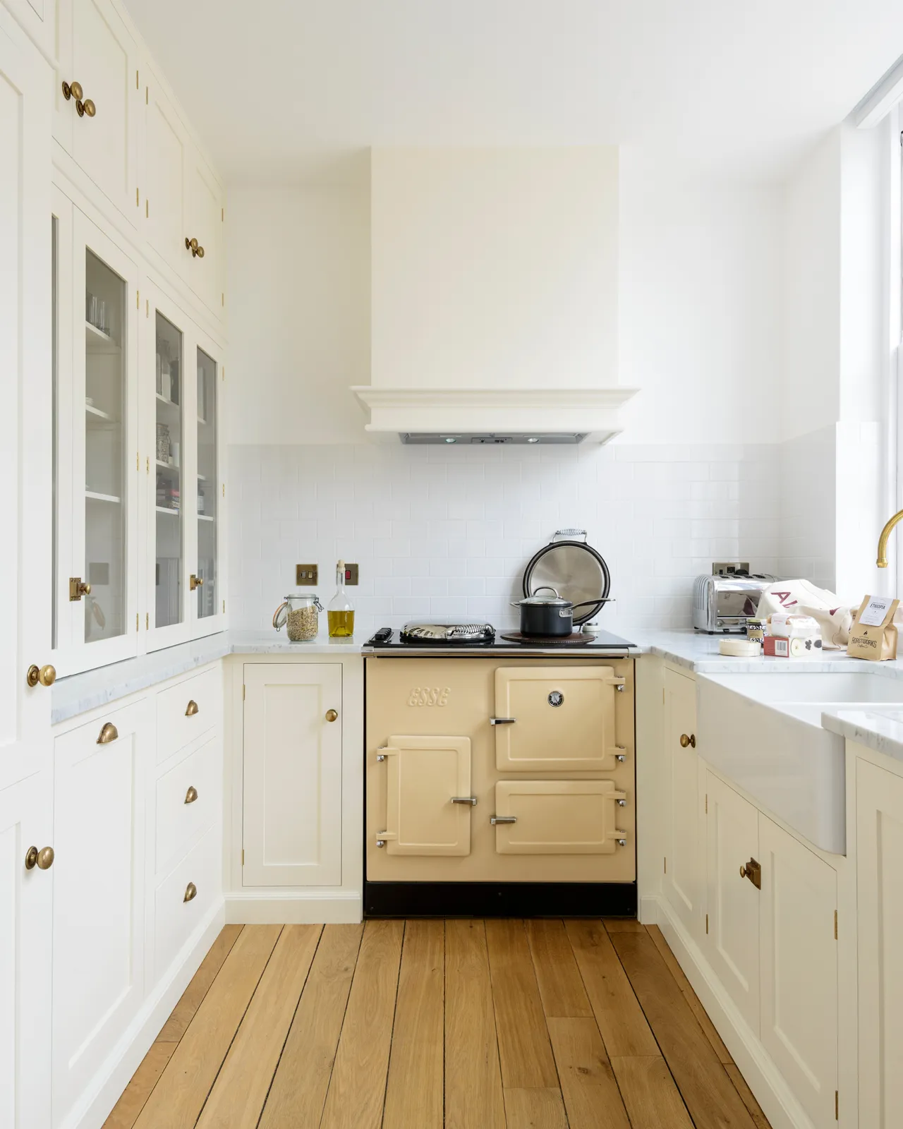 Looking into a small apartment kitchen with cream coloured Shaker-style cupboards around the room, with tall cupboards on the left and a sink below a window on right. On the back wall is a buttery Aga cooker with a small canopy extractor fan housing above.