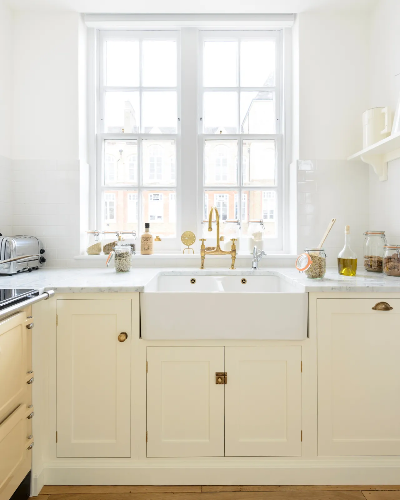 A straight-on view of a simple Shaker-style sink cupboard, painted in a cream colour with brass hardware, a ceramic Belfast sink and aged brass taps.