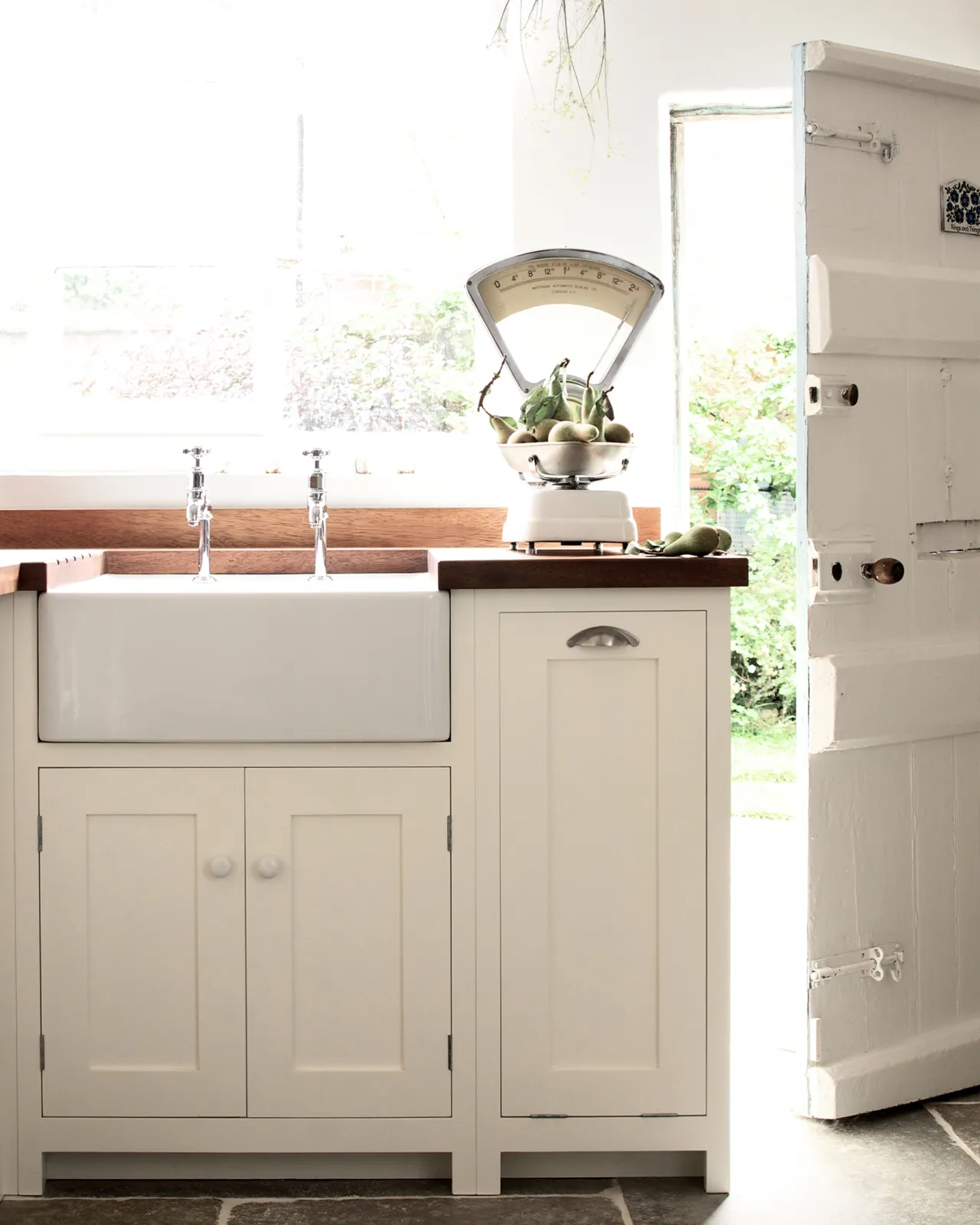 A straight-on view of a pair of traditional-style chrome bibcock taps with crosshead handles in a small cottage-style kitchen with a white ceramic Belfast sink and rich stave hardwood worktops.