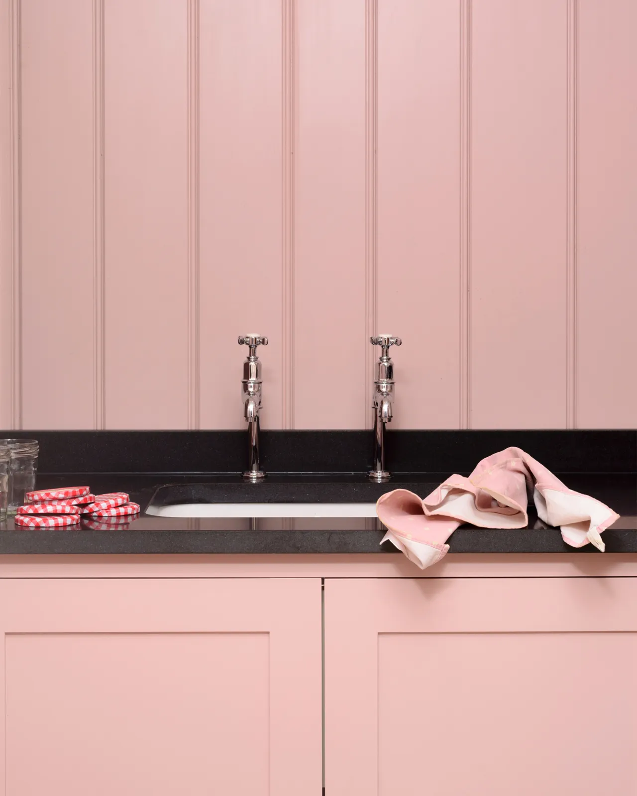 A straight-on view of a pair of traditional-style chrome bibcock taps with crosshead handles in a classic utility room with a black granite worktop, pink painted cupboards and tongue and groove panelling.