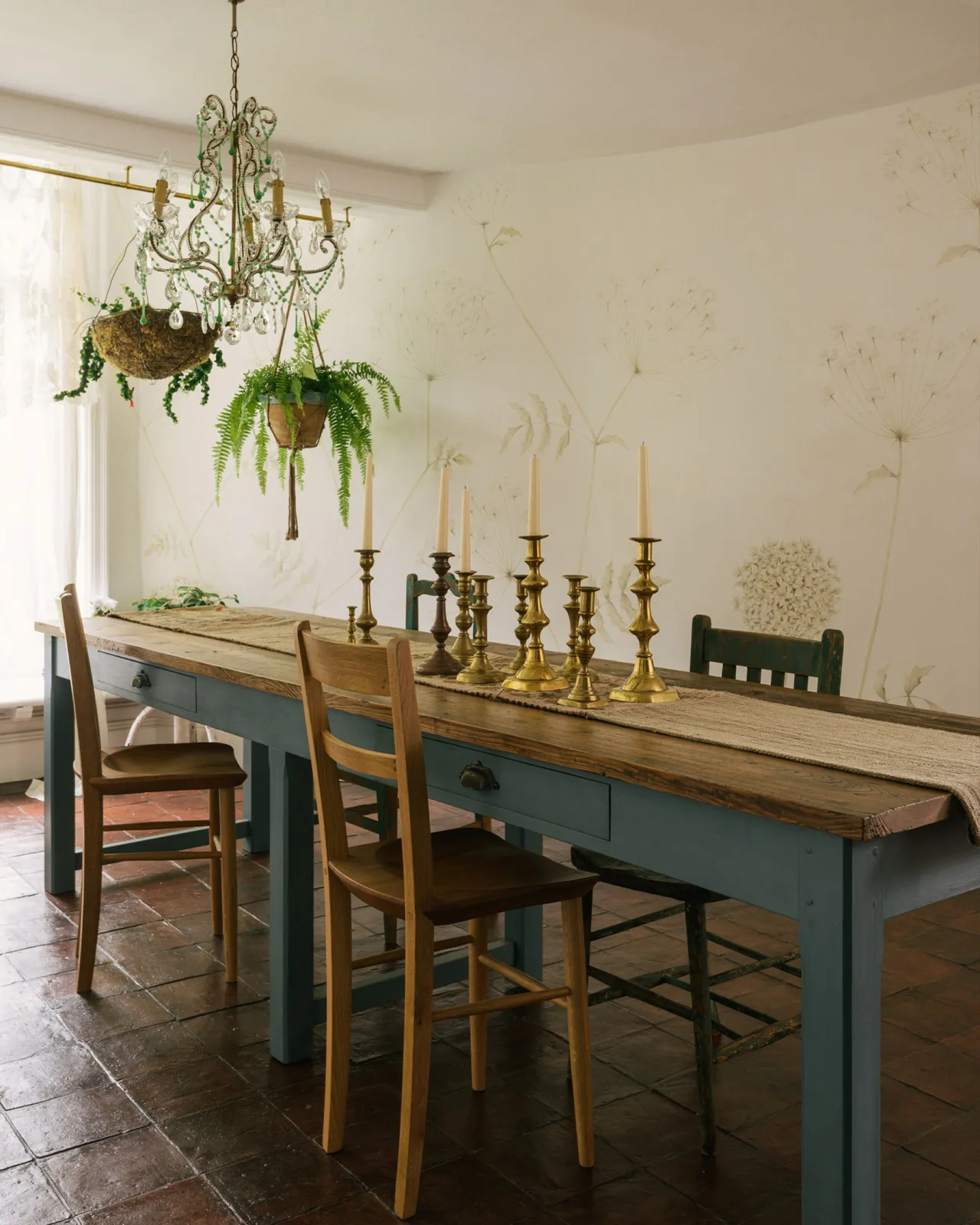 A long banquet-style dining table with simple blue-green painted legs and frame and rustic reclaimed pine boards for the tabletop. The room has vintage bohemian styling with mismatched chairs and hanging plants.