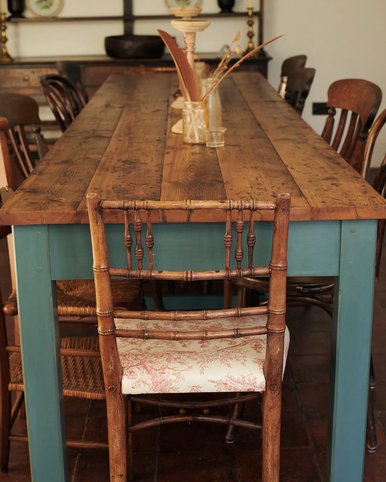An antique wooden chair at the end of a long banquet-style dining table with simple blue-green painted legs and frame and rustic reclaimed pine boards for the tabletop.