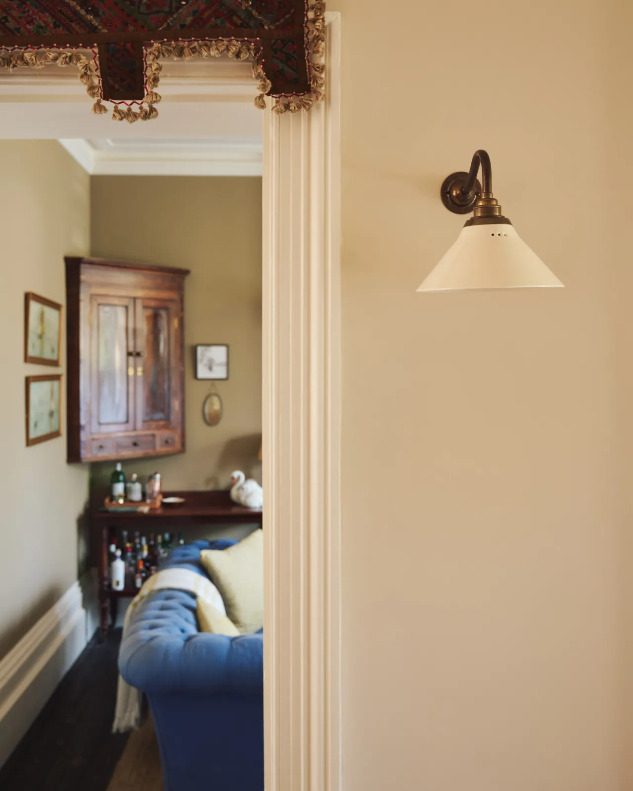 A straight-on view of a small wall light with an aged brass swan neck arm and a small cream glazed ceramic shade, fixed to a cream wall by an open doorway.