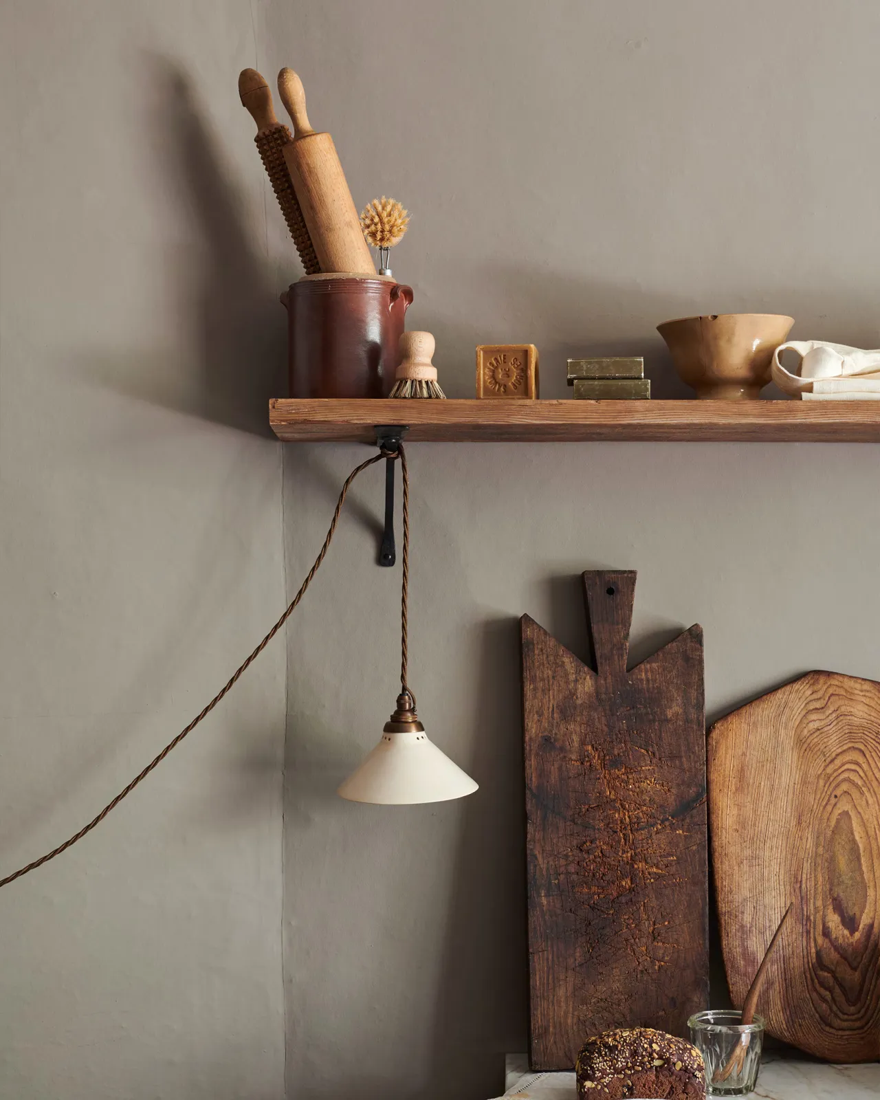 A straight-on view of a small plug-in light with a cream glazed shade, brown twisted fabric flex cord and brass fittings, hanging from an iron shelf bracket under a rustic pine shelf.