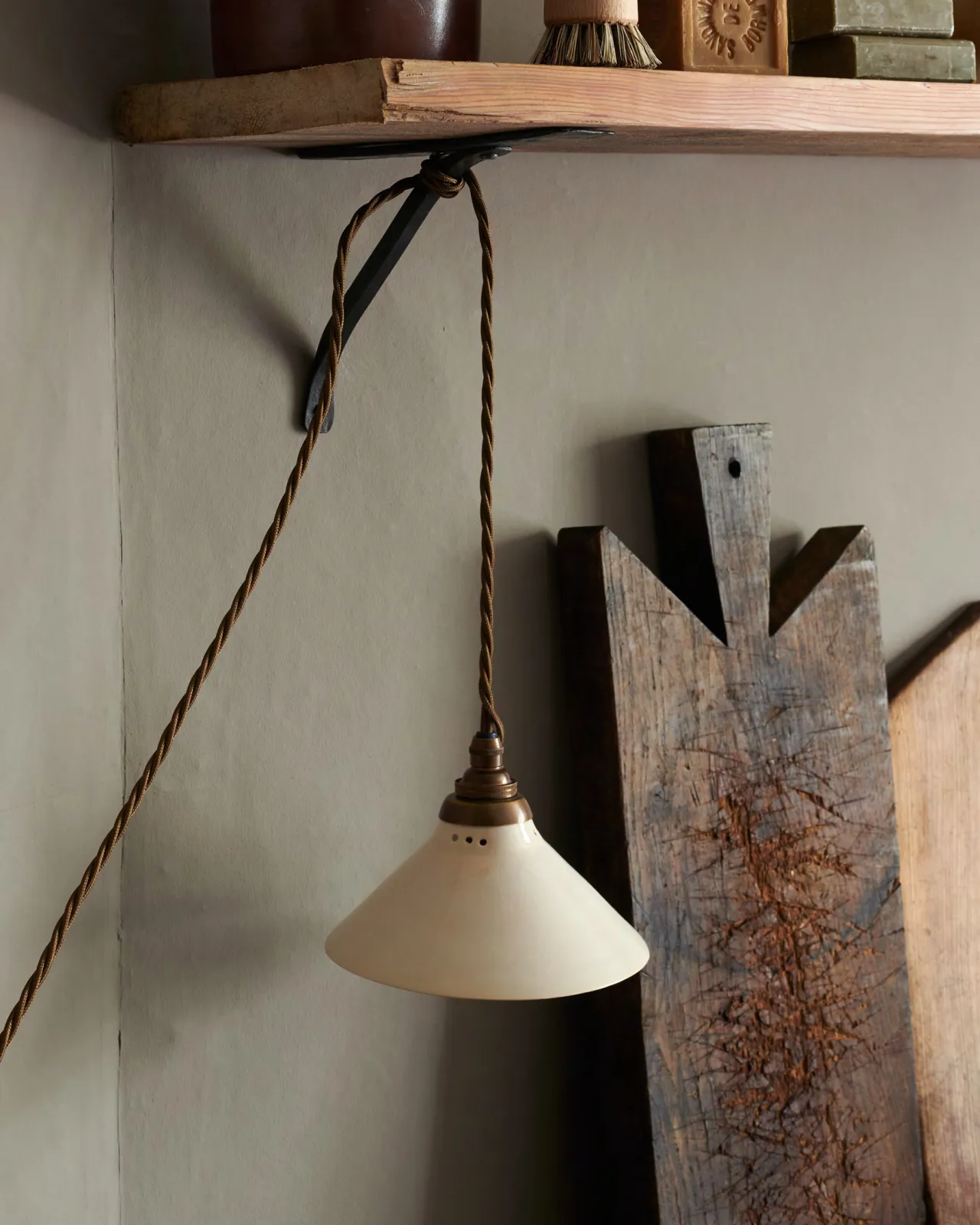 A close-up oblique view of a small plug-in light with a cream glazed shade, brown twisted fabric flex cord and brass fittings, hanging from an iron shelf bracket under a rustic pine shelf.