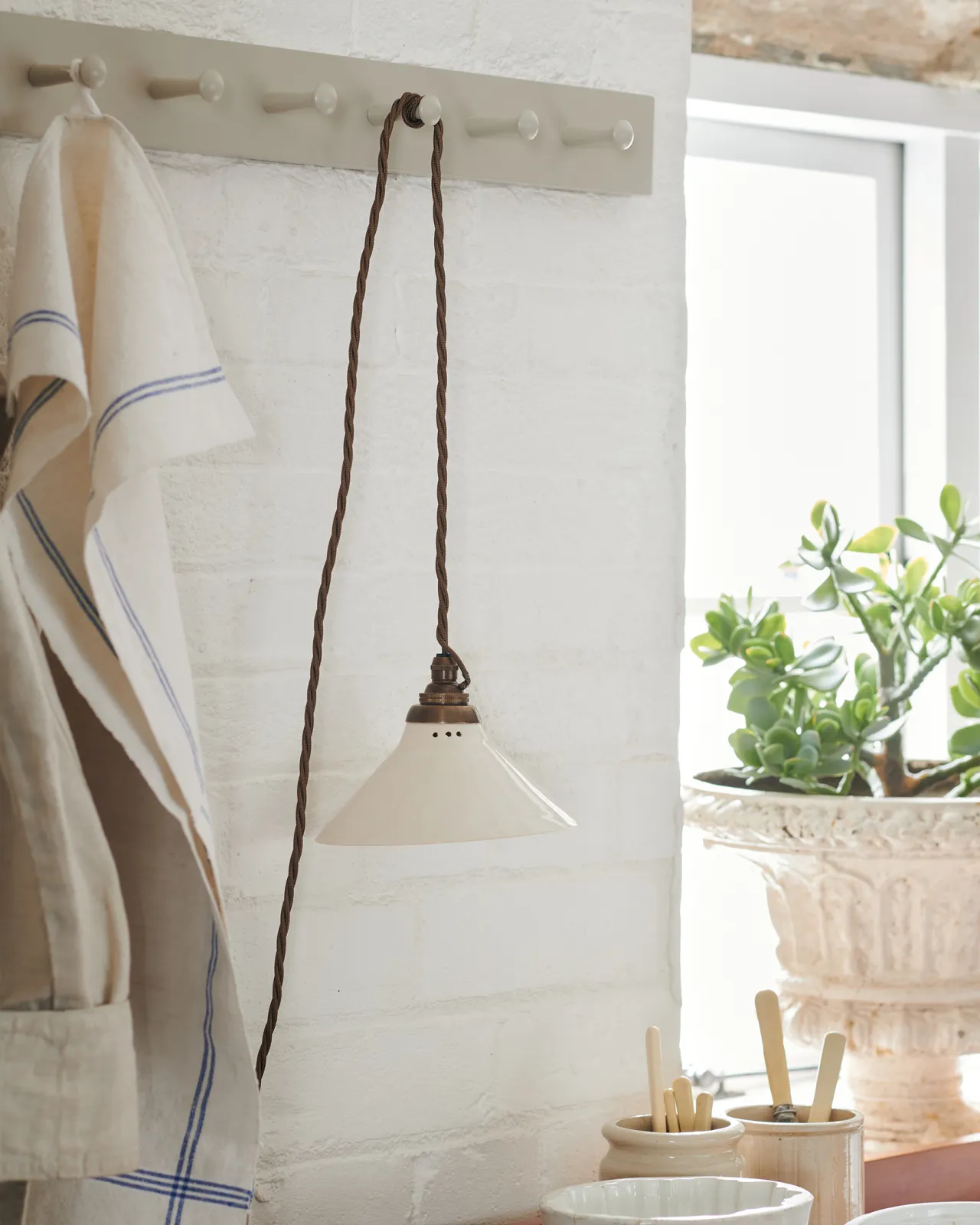 A close-up oblique view of a small plug-in light with a cream glazed shade, brown twisted fabric flex cord and brass fittings, hanging from painted pegs above a kitchen worktop.