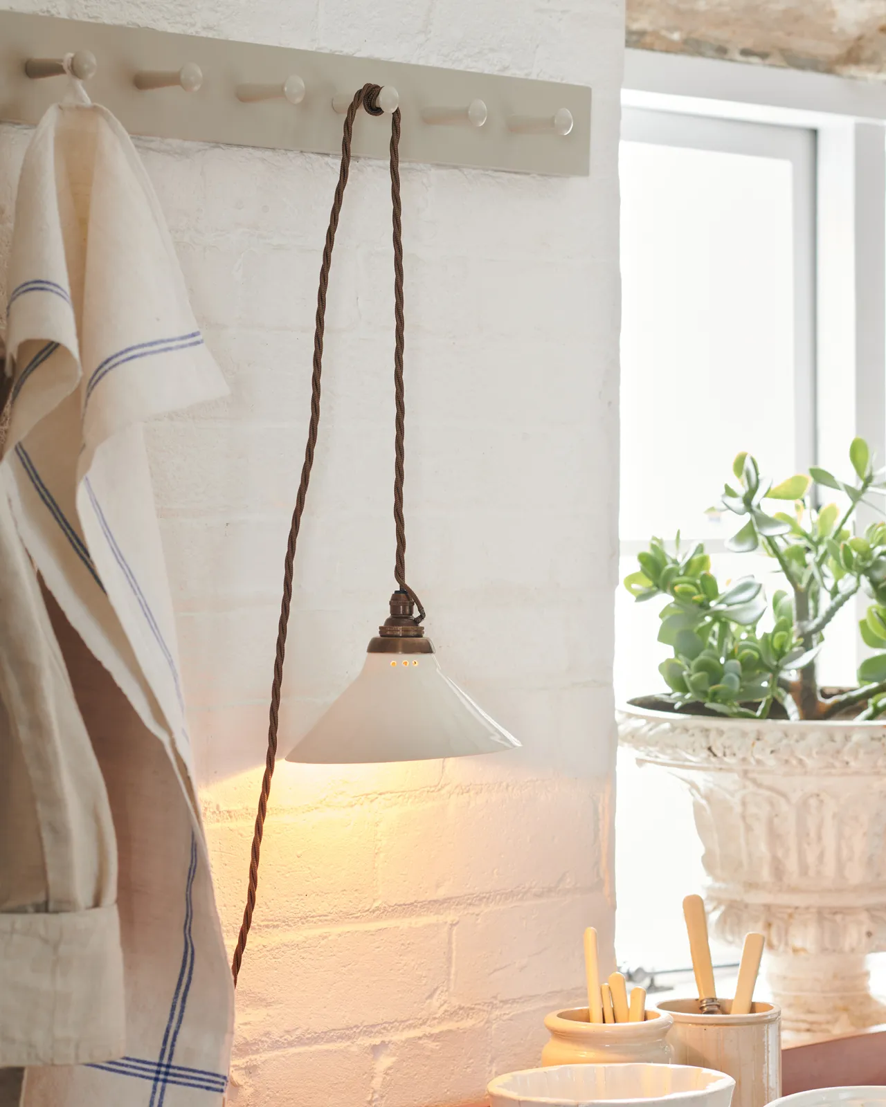 A close-up oblique view of a small plug-in light with a cream glazed shade, brown twisted fabric flex cord and brass fittings, hanging from painted pegs above a kitchen worktop. The light is on.