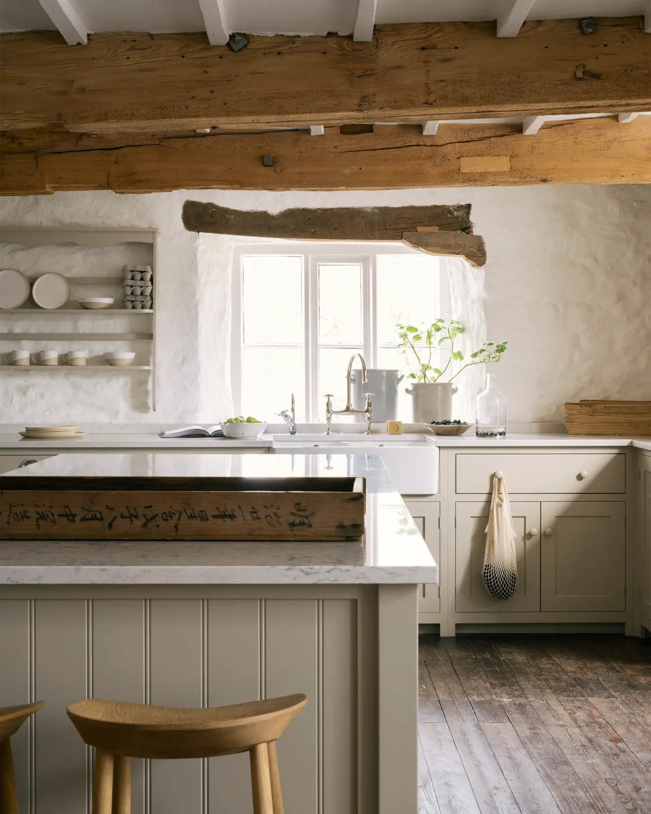 A large cottage kitchen with creamy mushroom coloured Shaker-style kitchen cupboards, dark worn floorboards and large wooden ceiling beams.