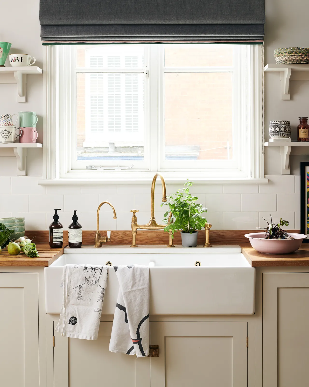 A close-up view of a wide ceramic Belfast sink in a Shaker-style kitchen with creamy mushroom painted cupboards, aged brass taps and a light hardwood worktop.