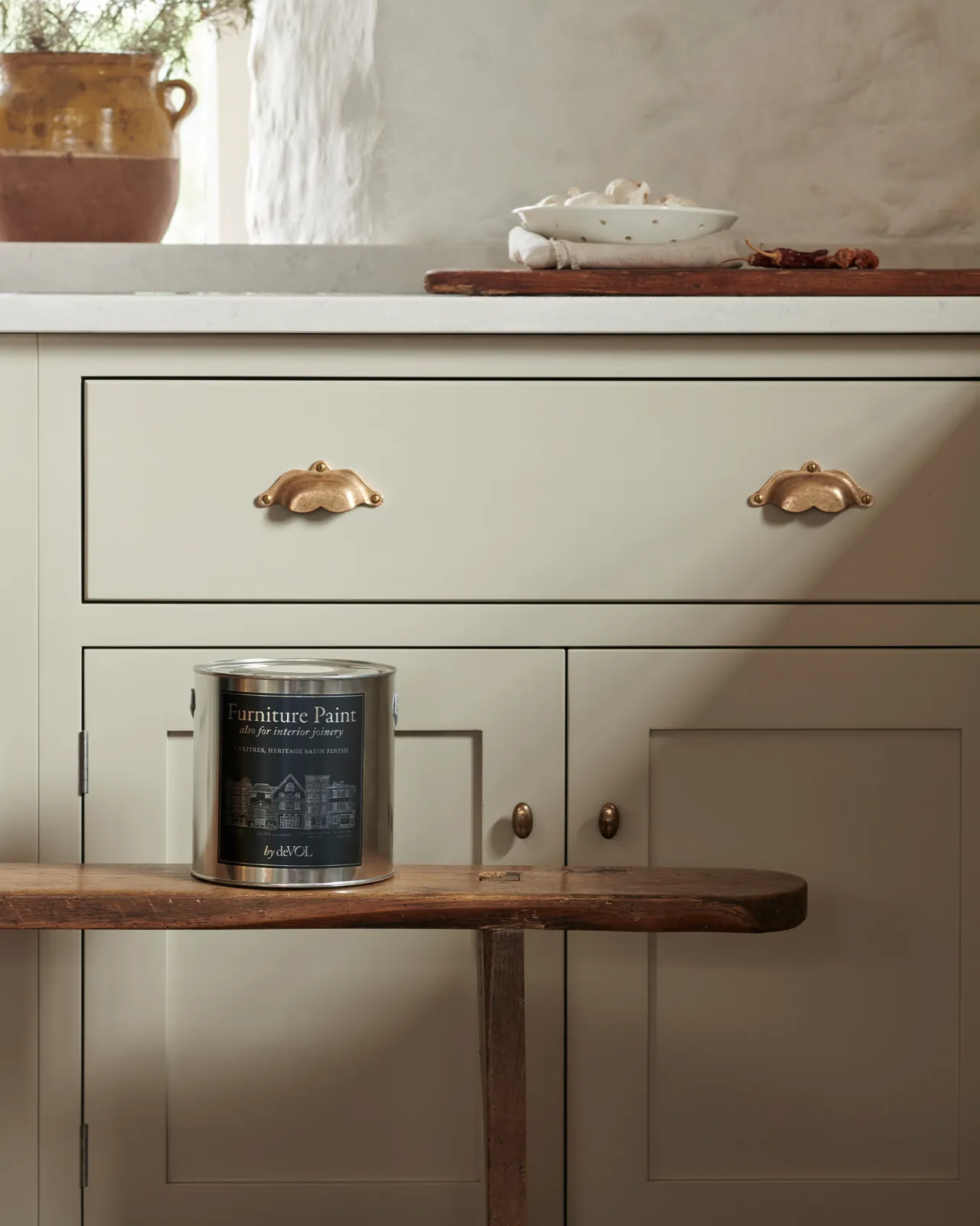 A silver paint tin with a blue label on an antique bench, in front of a mushroom coloured Shaker-style door/drawer base cabinet with ornate bronze pull handles. 