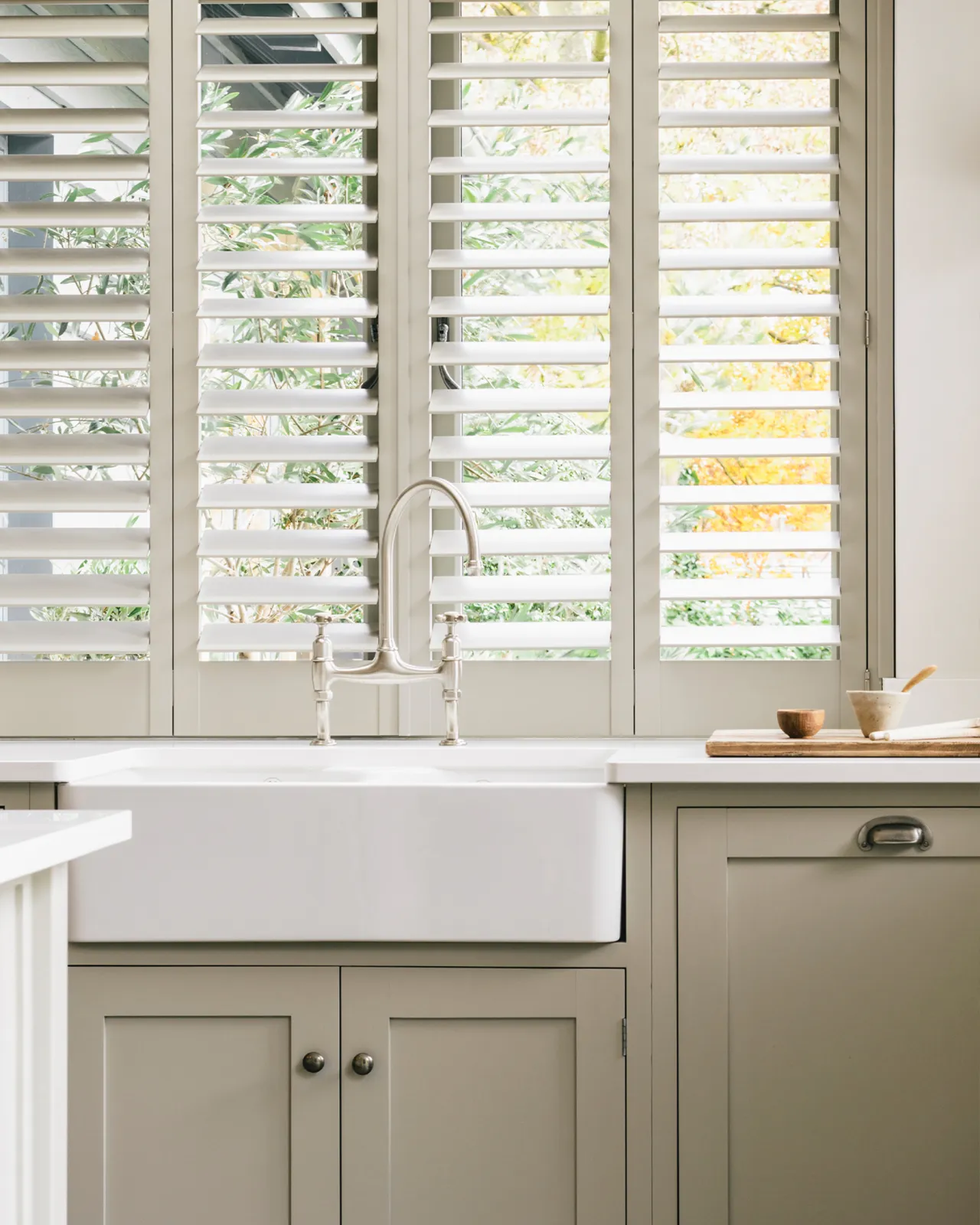 A straight-on view of a traditional-style nickel mixer tap with crosshead handles in a simple cream-coloured Shaker kitchen with louvred shutters behind.