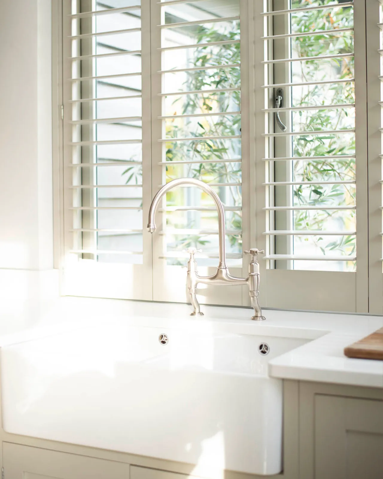 An oblique view of a traditional-style nickel mixer tap with crosshead handles in a simple cream-coloured Shaker kitchen with louvred shutters behind.