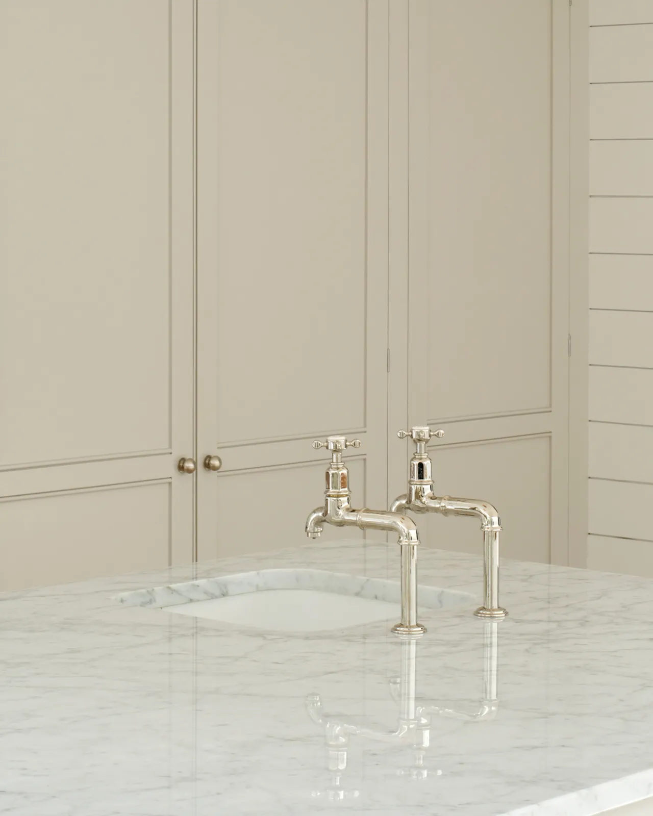 A pair of traditional-style nickel bibcock taps with crosshead handles on a polished Carrara marble worktop, creamy coloured kitchen cupboards can be seen in the background.