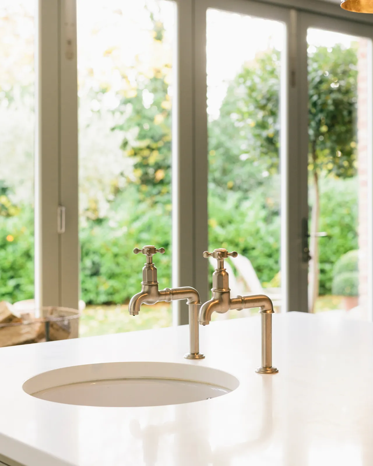 A close-up view of a pair of traditional-style nickel bibcock taps with crosshead handles over a round undermounted sink.