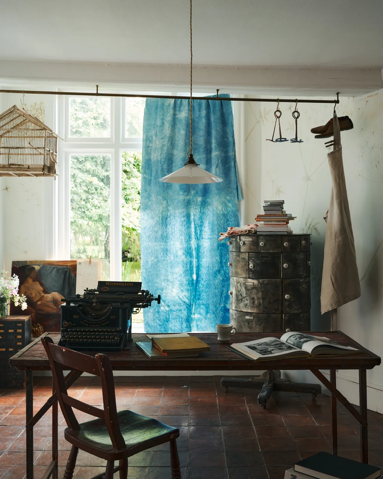 A straight-on view of a classic pendant light with a milk glass light shade, brass fittings and a cream twisted fabric flex cord, hanging above a typewriter and scraps of paper.