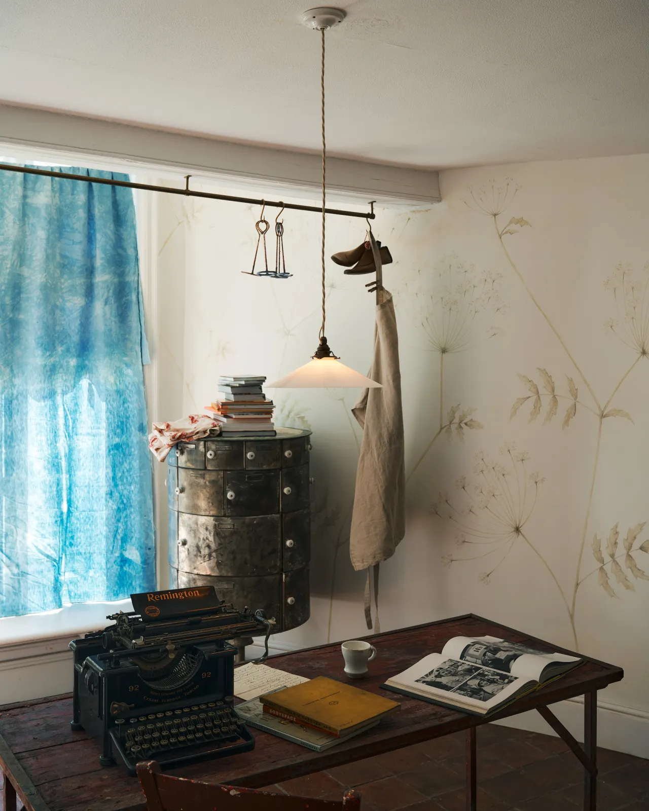 An oblique view of a classic pendant light with a milk glass light shade, brass fittings and a cream twisted fabric flex cord, hanging above a typewriter and scraps of paper.