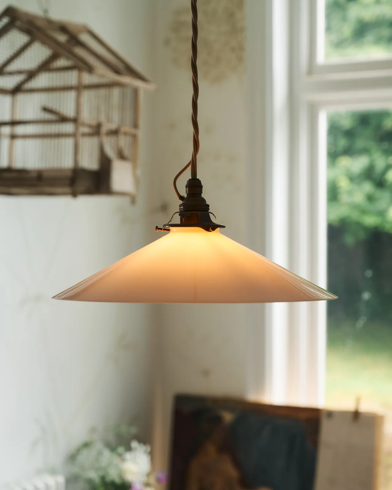 A close-up view of a classic pendant light with a milk glass light shade, brass fittings and a cream twisted fabric flex cord. The light is on, showing the warm red glow of the translucent surface.