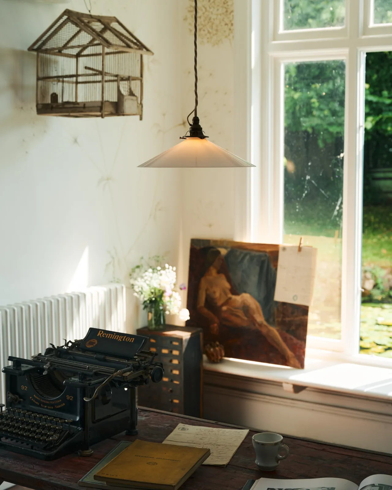 An oblique view of a classic pendant light with a milk glass light shade, brass fittings and a cream twisted fabric flex cord, hanging above a typewriter and scraps of paper by an old oil painting and a large window.