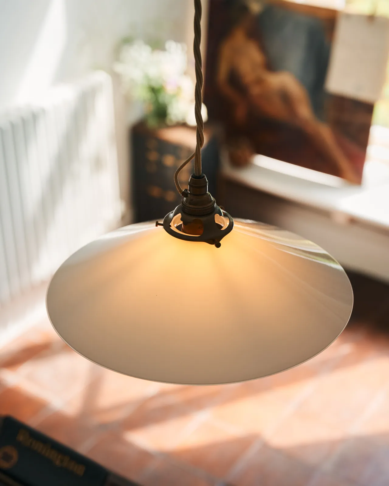 A close-up oblique view of a classic pendant light with a milk glass light shade, brass fittings and a cream twisted fabric flex cord. The light is on, showing the warm red glow of the translucent surface.