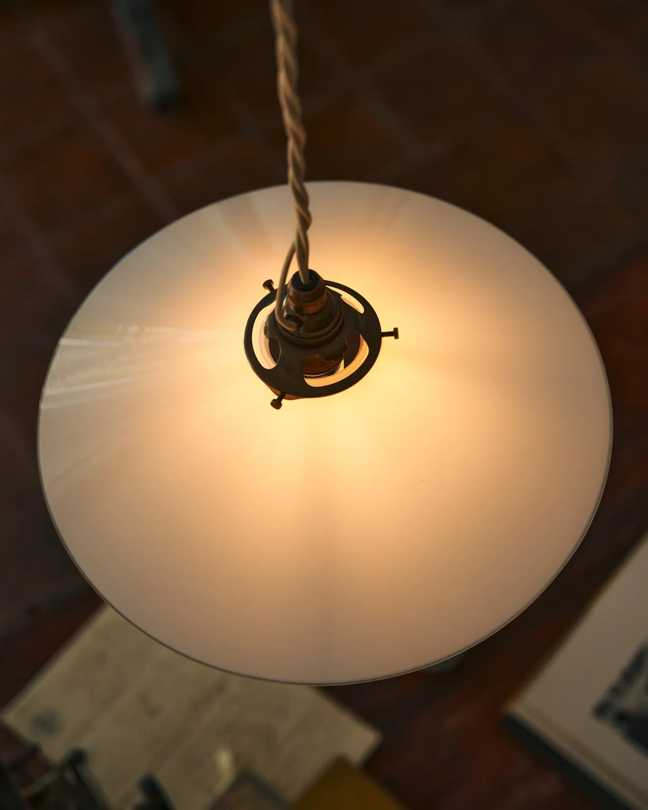 A close-up overhead view of a classic pendant light with a milk glass light shade, brass fittings and a cream twisted fabric flex cord. The light is on, showing the warm red glow of the translucent surface.