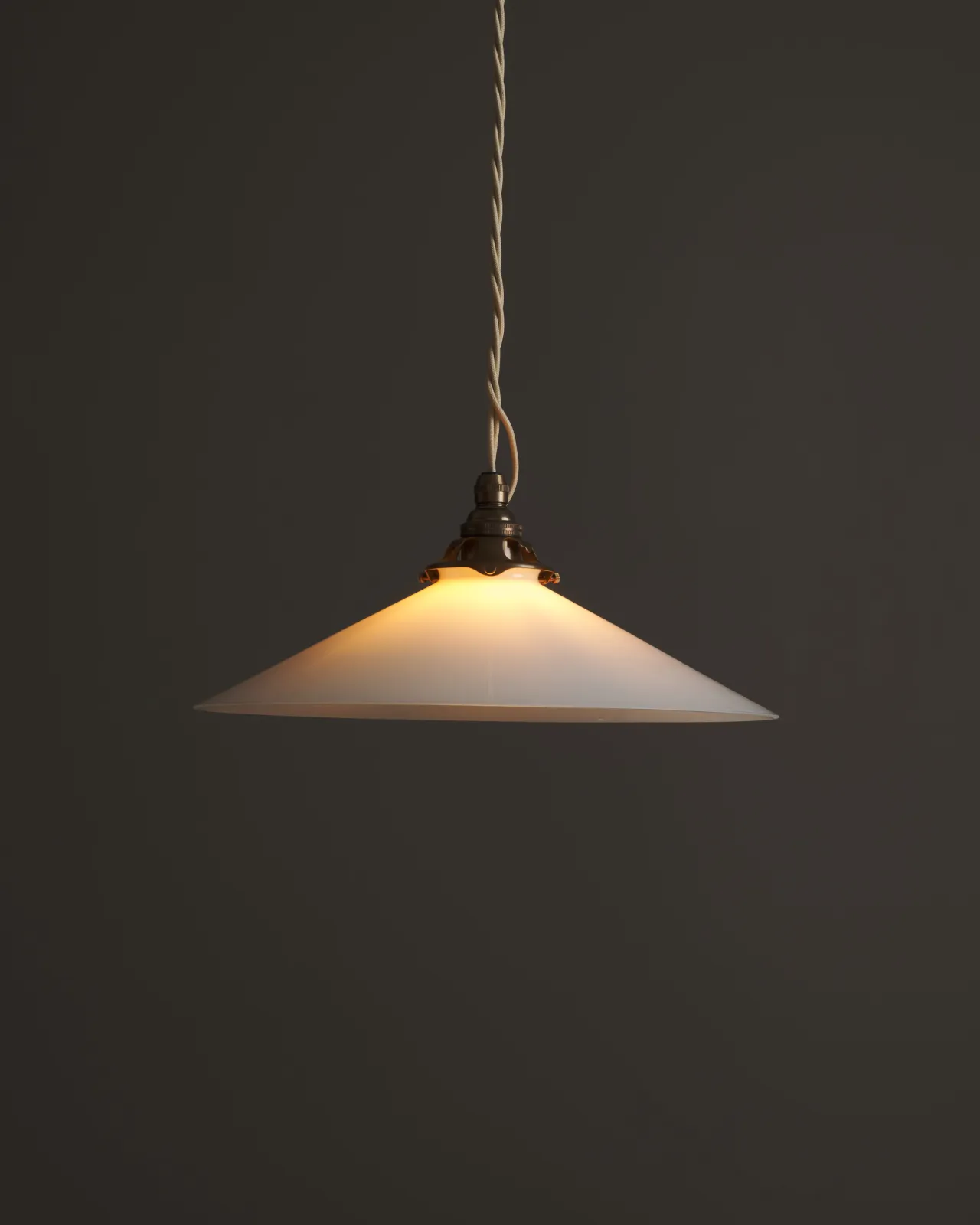 A classic pendant light with a milk glass light shade, an aged brass E27 lampholder with a decorative gallery and cream twisted fabric flex cord. The light is on, showing the warm red glow of the translucent surface.