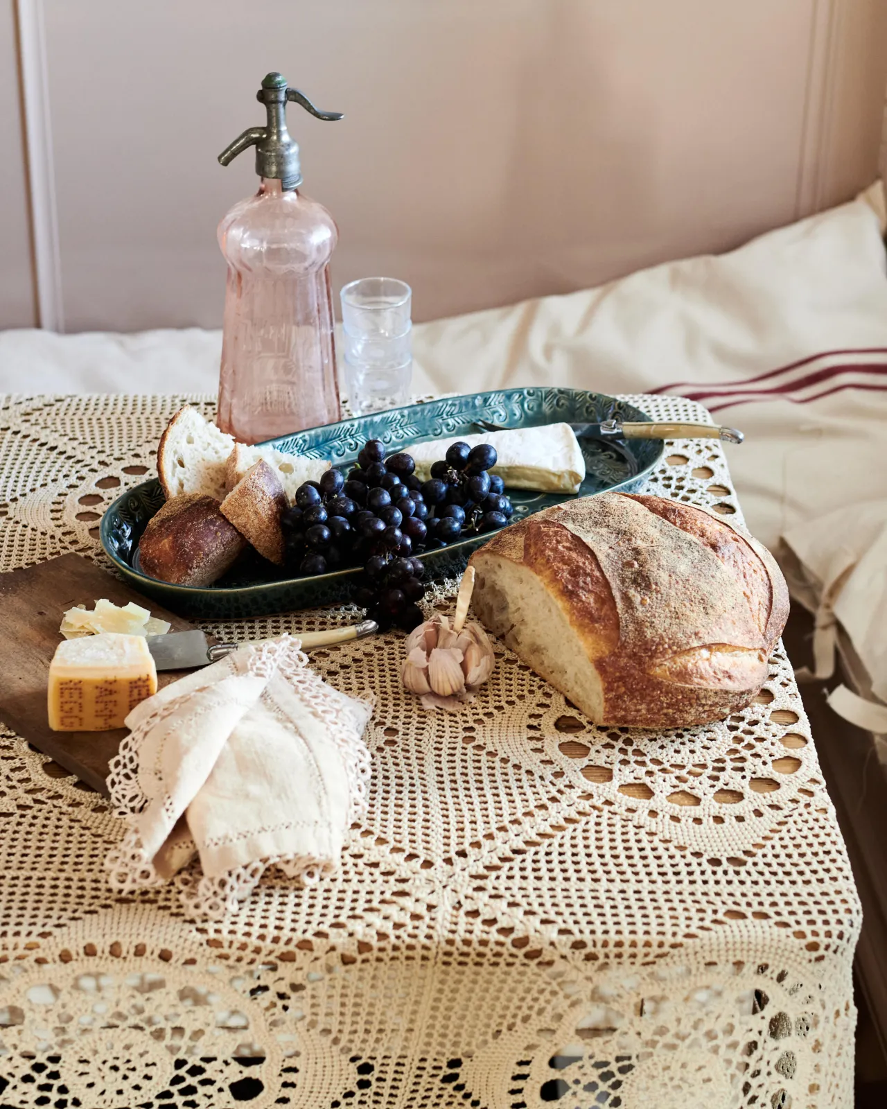 An oval-shaped handmade bohemian-style blue ceramic platter, with black grapes, cheeses and homemade bread on a beautiful crochet lace tablecloth.