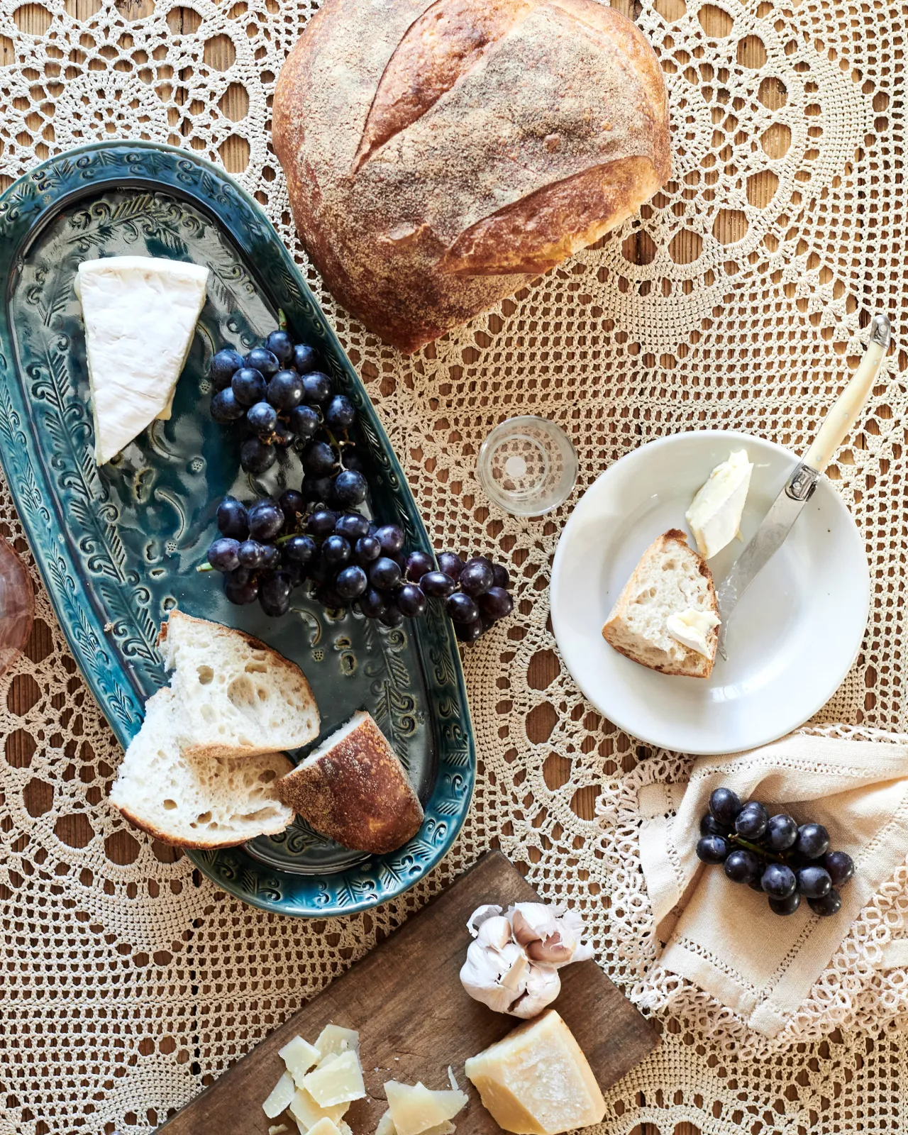 An overhead view of an oval-shaped handmade bohemian-style blue ceramic platter, with black grapes, cheeses and homemade bread on a beautiful crochet lace tablecloth.