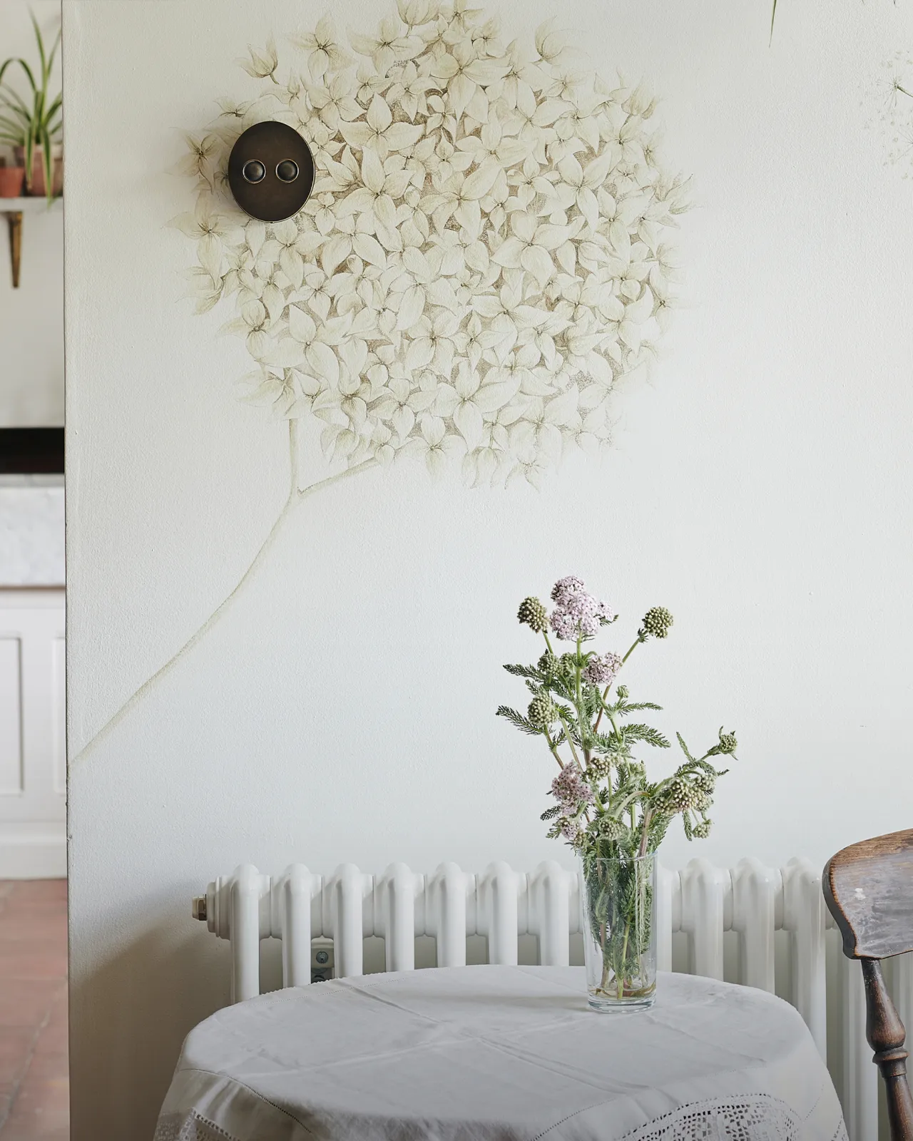 A straight-on view of a 2 gang oval-shaped dimmer switch in a dark oxidised brass finish on a white wall with a large mural of an allium flower head. A small round table with a white linen tablecloth and a vase of flowers is in the foreground.