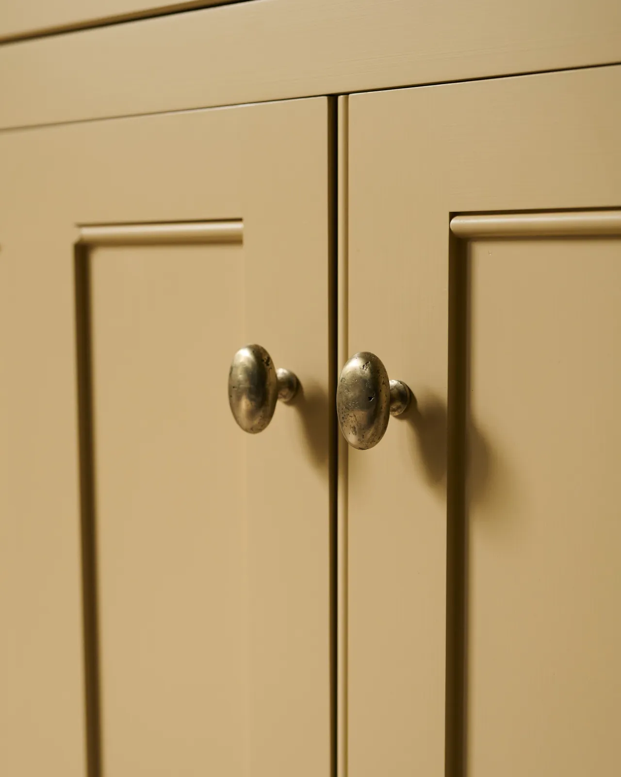An oblique view of a pair of oval-shaped knobs in an aged brass finish, fitted to a yellow painted Georgian-style kitchen cupboard.