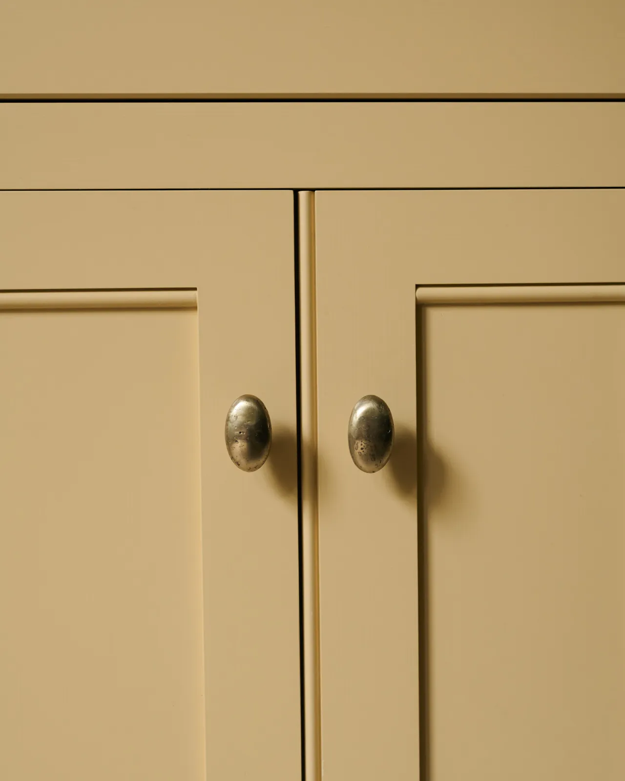 A straight-on view of a pair of oval-shaped knobs in an aged brass finish, fitted to a yellow painted Georgian-style kitchen cupboard.