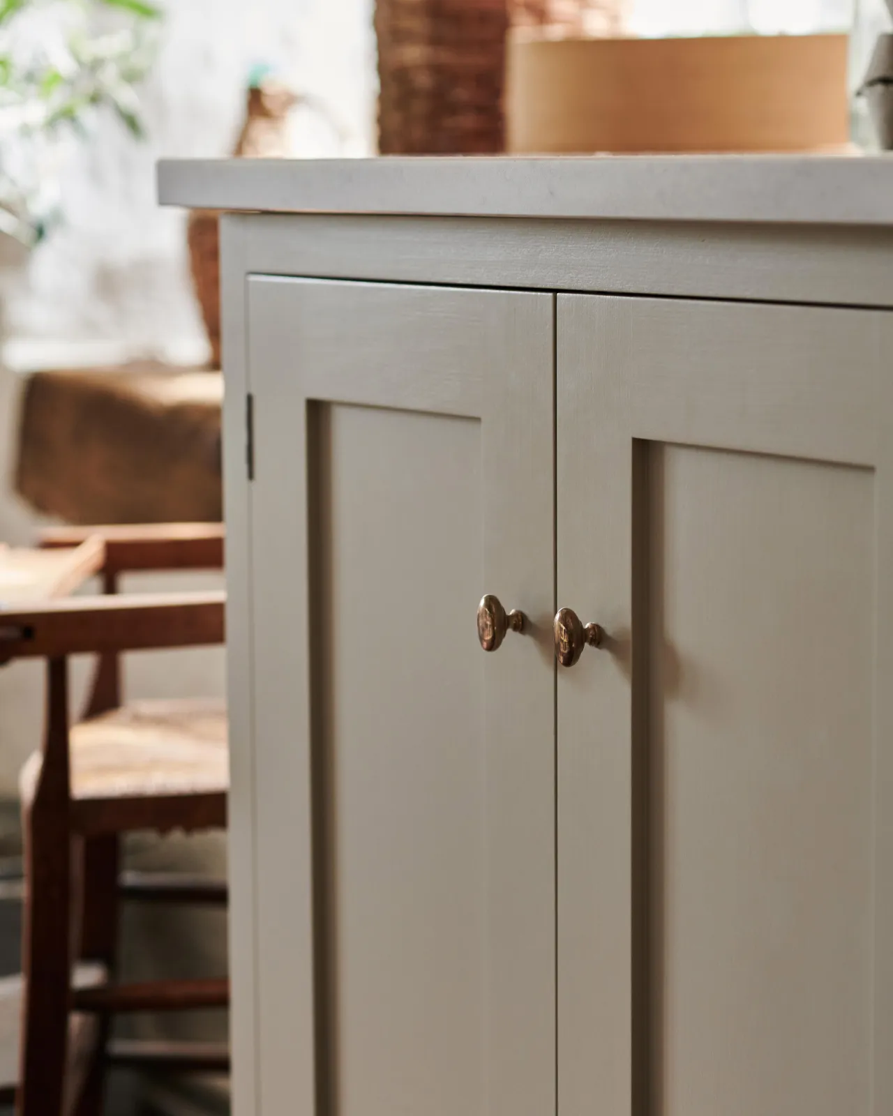 An oblique view of a pair of oval-shaped knobs in a warm burnished brass finish, fitted to a mushroom painted Shaker-style kitchen cupboard.