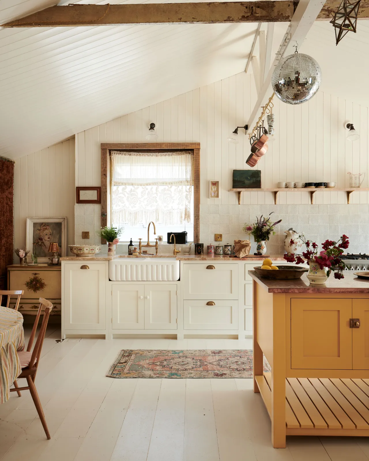 A bohemian kitchen with cream coloured Shaker-style cupboards, floorboards and walls. A disco-ball hangs from a ceiling joist above a warm yellow coloured freestanding prep table with an aged copper worktop.