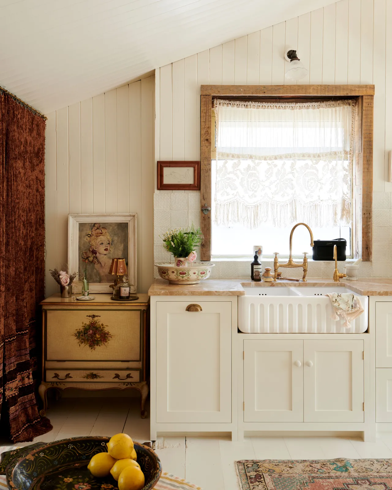 A bohemian kitchen with cream coloured Shaker-style cupboards, floorboards and walls. The room is decorated with an assortment of vintage pieces of furniture, paintings and fabrics.