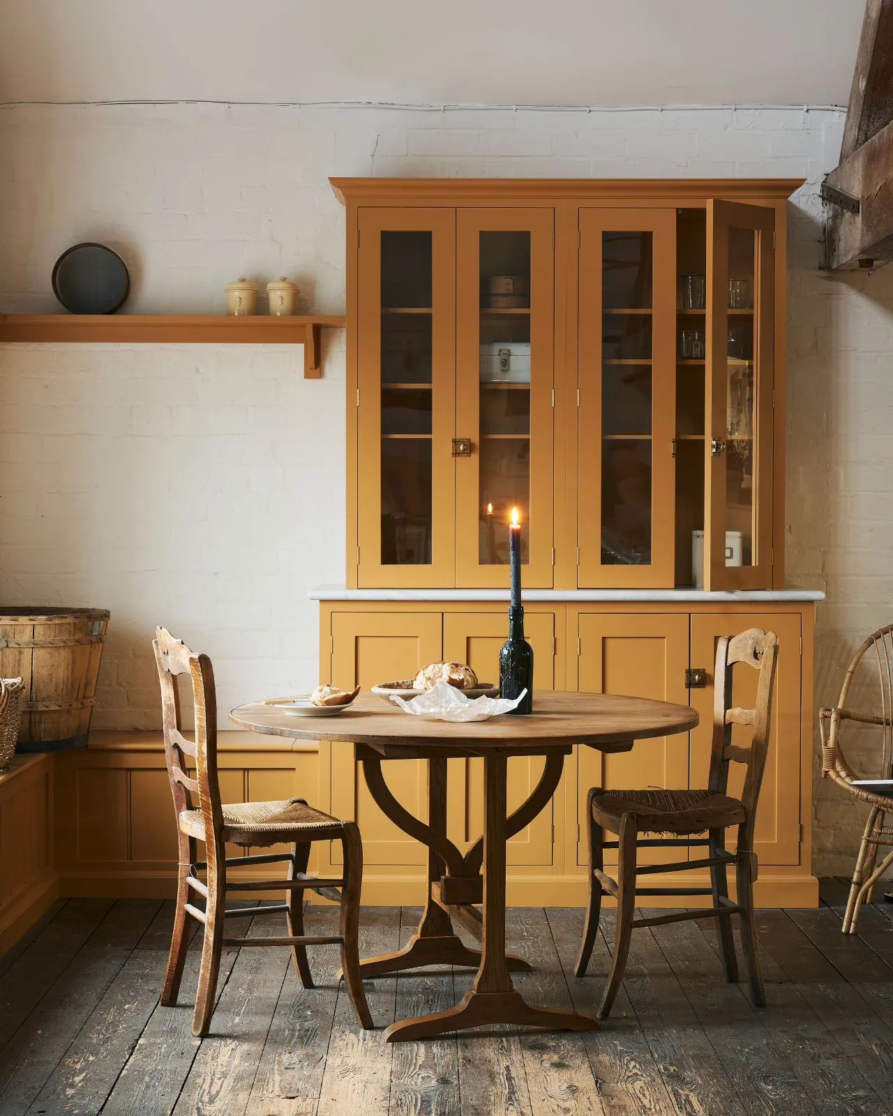 A straight-on view of a round pine dining table with vintage wooden chairs, in a rustic cottage kitchen with yellow painted Shaker-style cabinetry, dark worn floorboards, white brick walls and wooden beams.