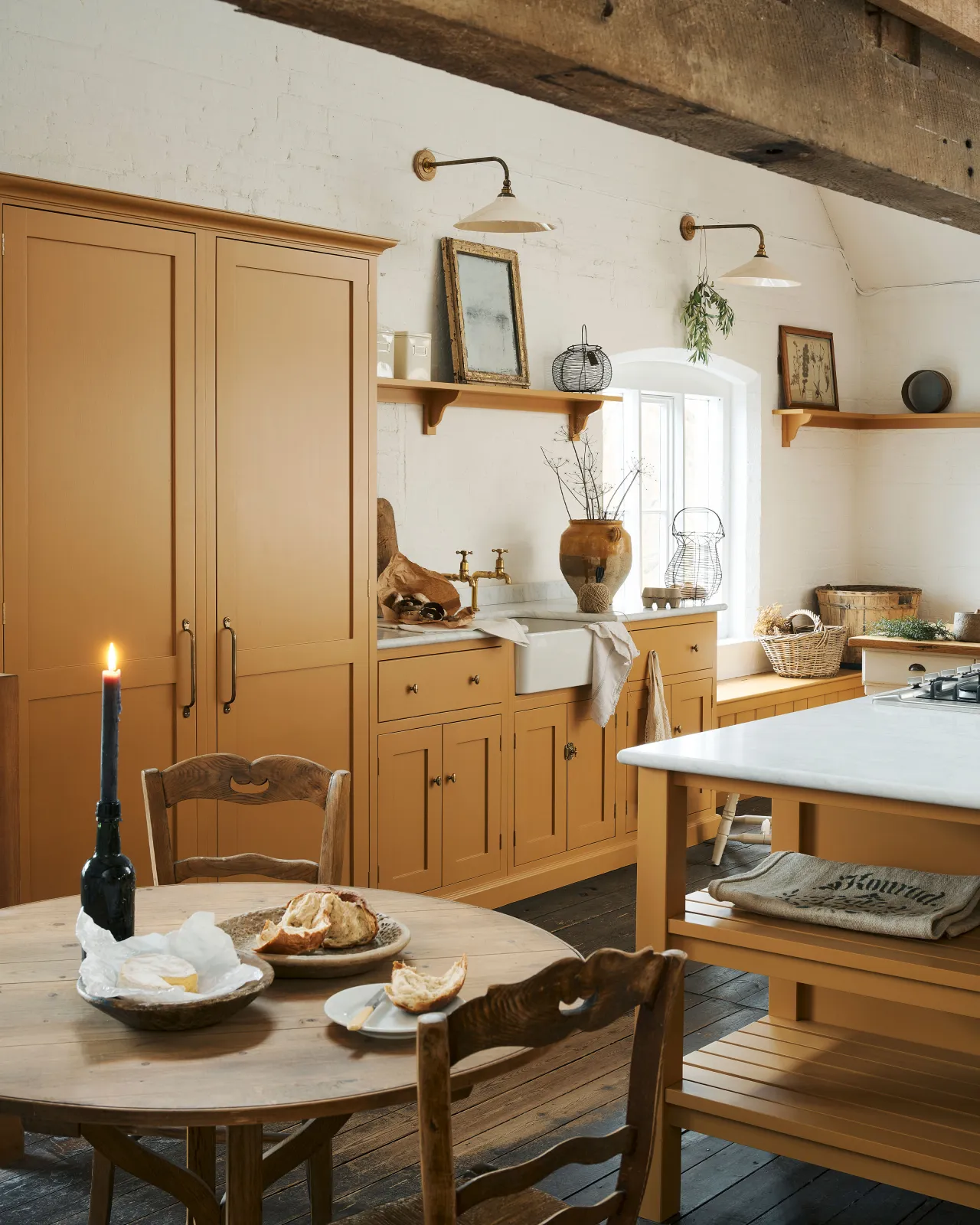 An oblique view of a yellow painted Shaker-style kitchen in a rustic cottage kitchen with dark worn floorboards, white brick walls and wooden beams.