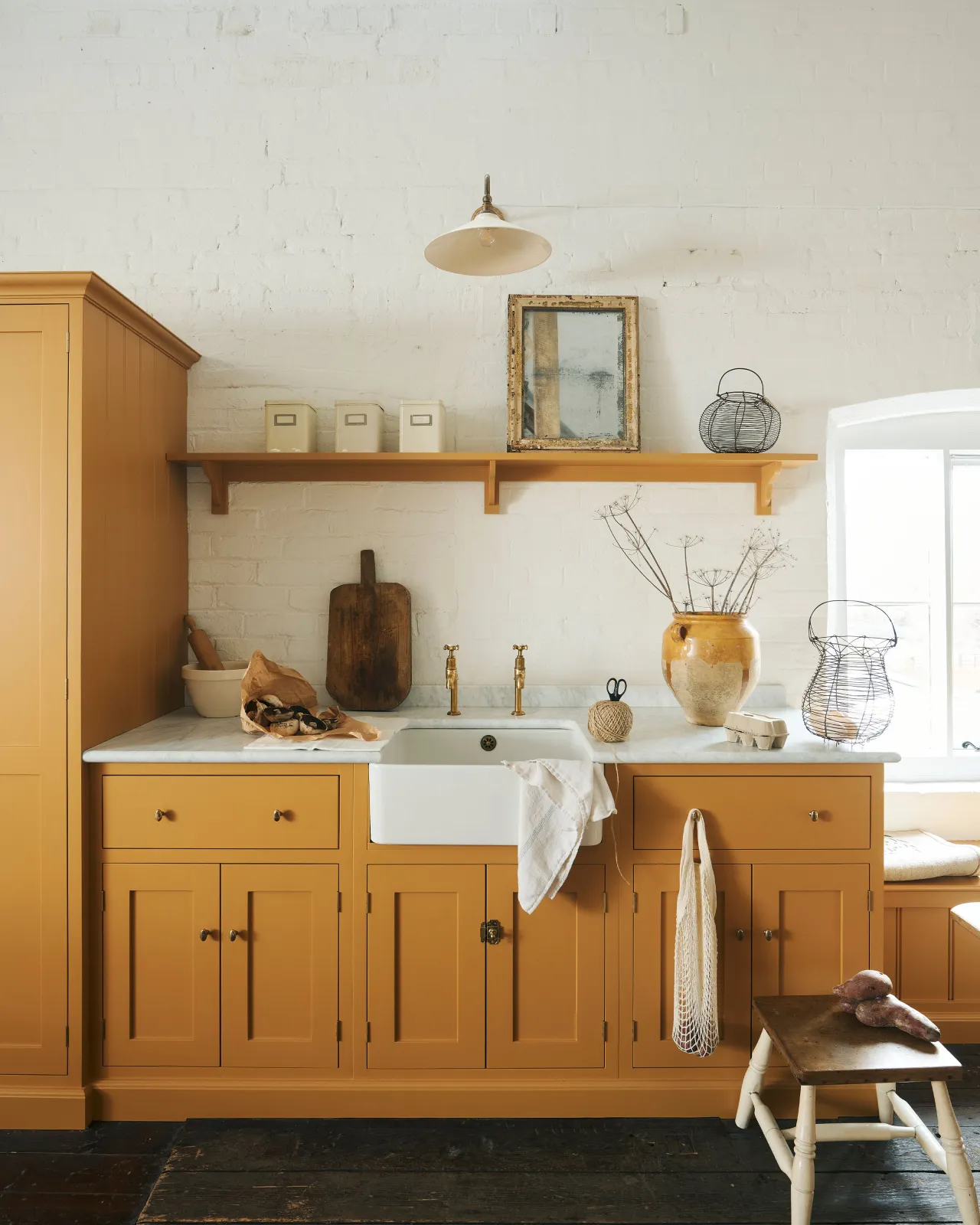 A straight-on view of a yellow painted Shaker-style kitchen in a rustic cottage kitchen with dark worn floorboards, white brick walls and wooden beams.