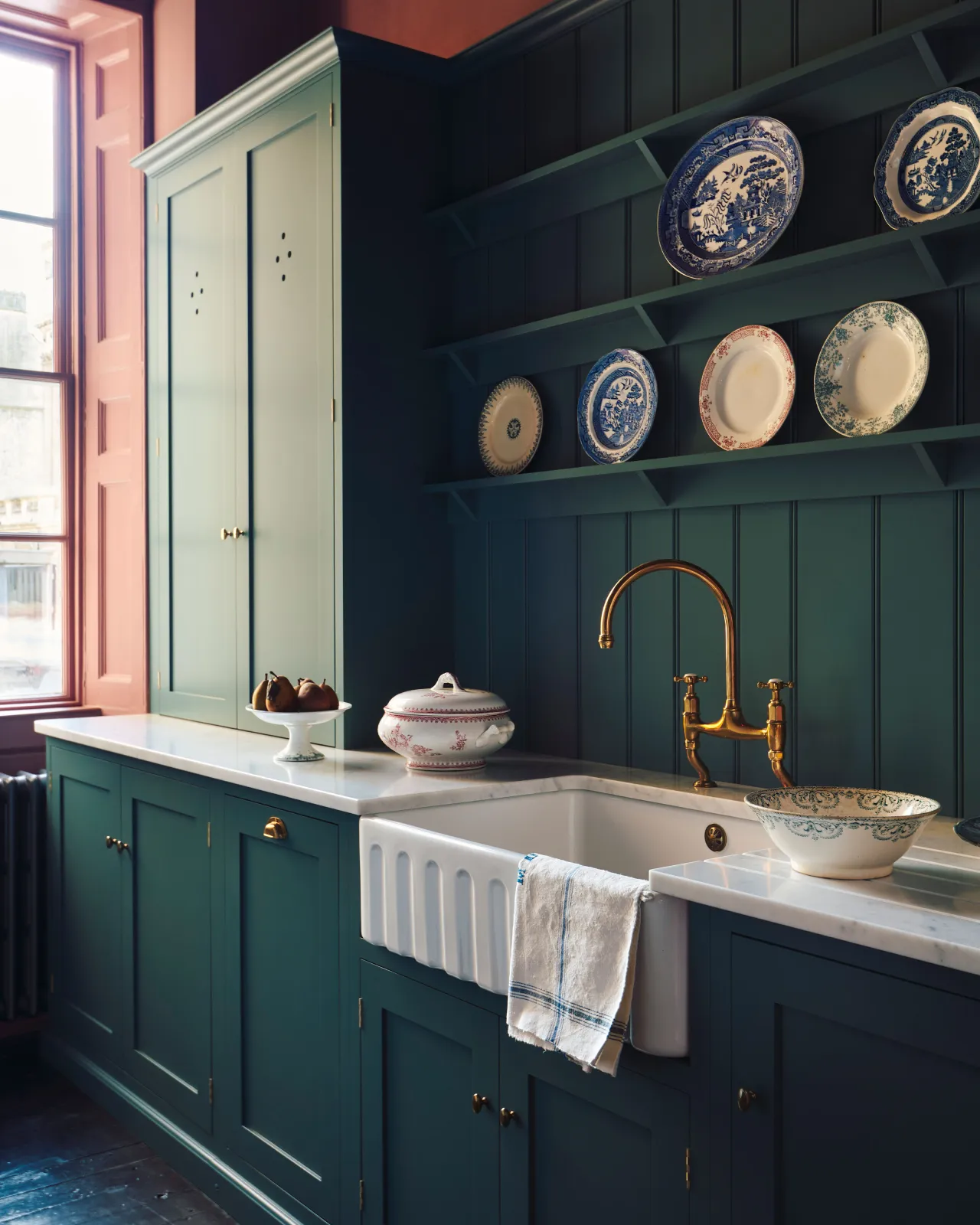 An oblique view of a classically-styled kitchen sink with green-blue painted Shaker-style cupboards, tongue and groove wall panelling, shelving full of vintage crockery and wine red coloured walls.